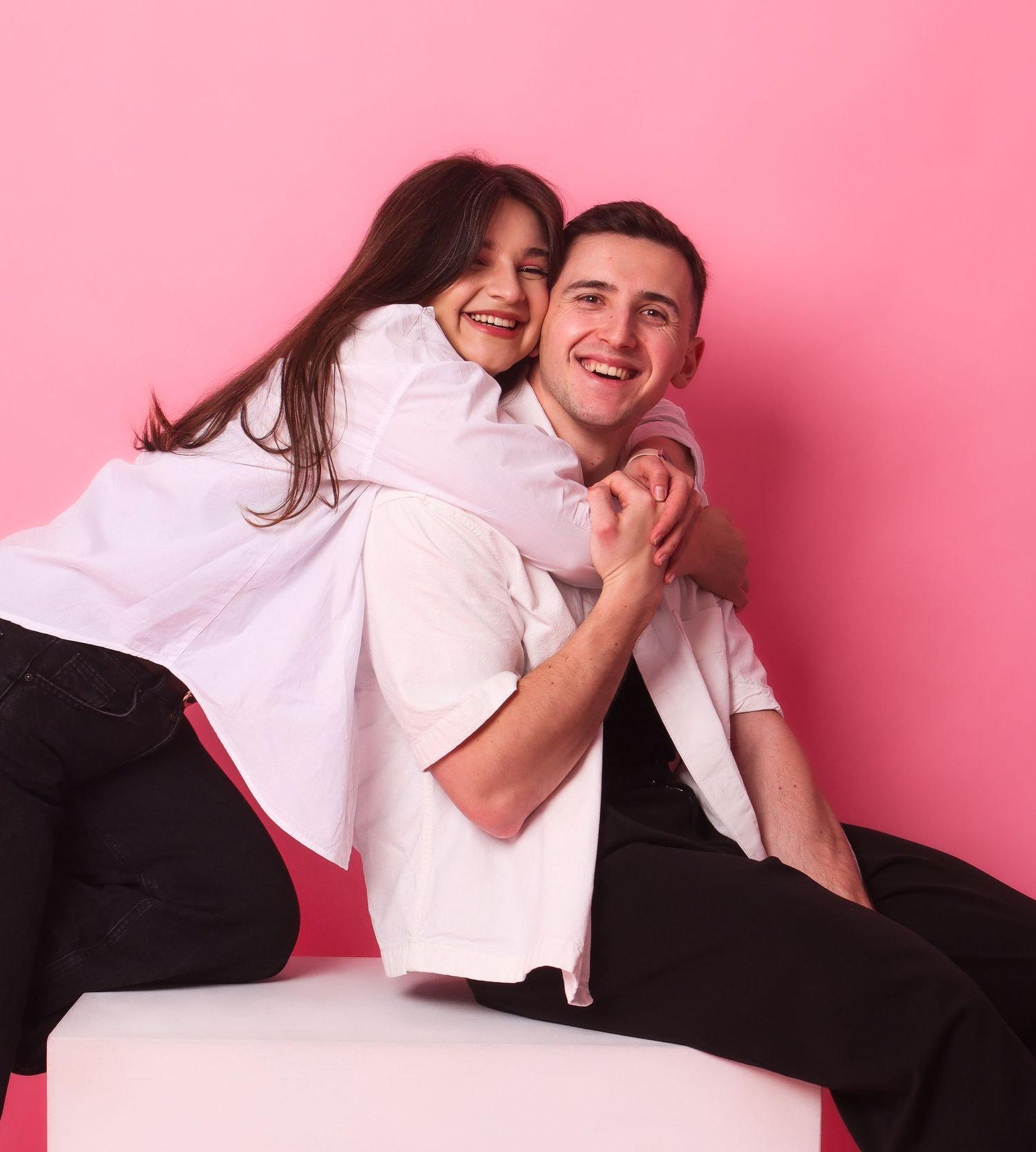 A happy couple laughs and cuddles on a white cube against a bright pink background.
