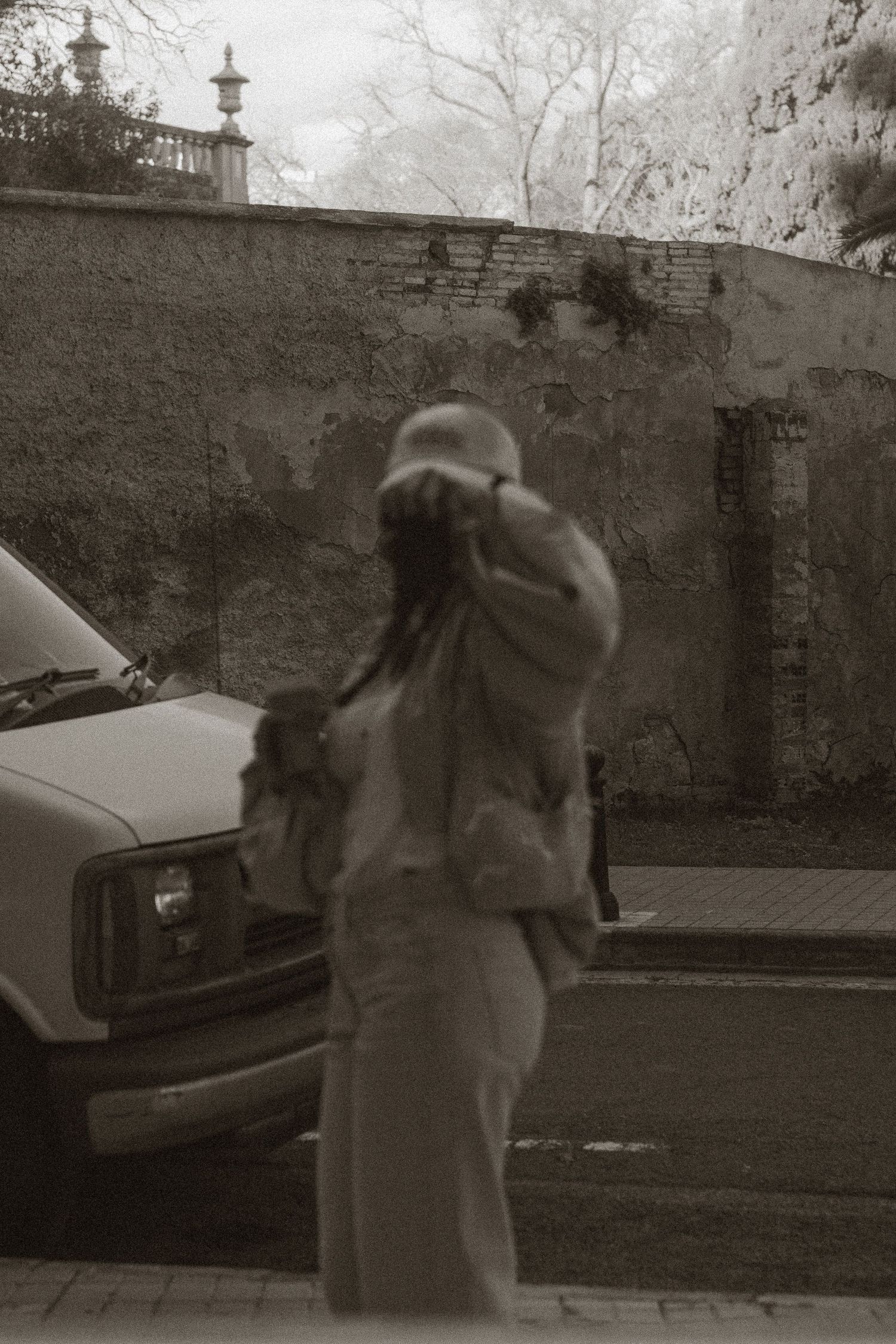 Person photographing outdoors near a vintage car and brick wall in black and white.