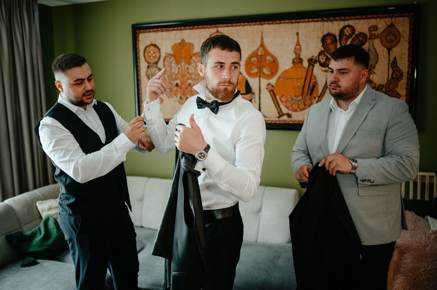 Three groomsmen in formal wear putting on suit jackets together in a decorated hotel room.