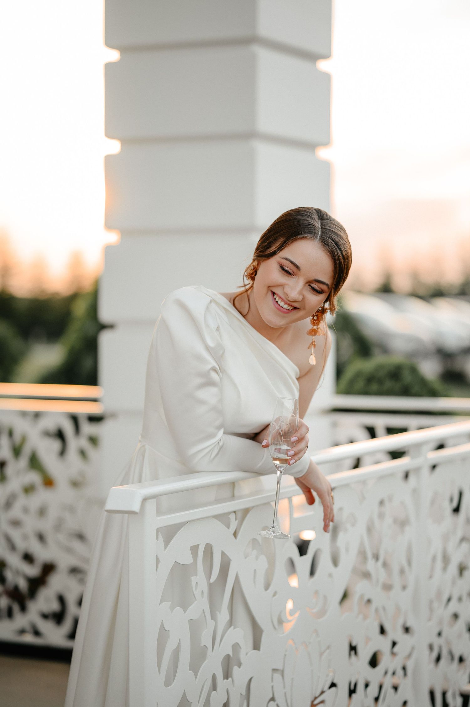 Joyful bride in white asymmetric gown laughs while leaning on ornate white balcony railing at sunset.