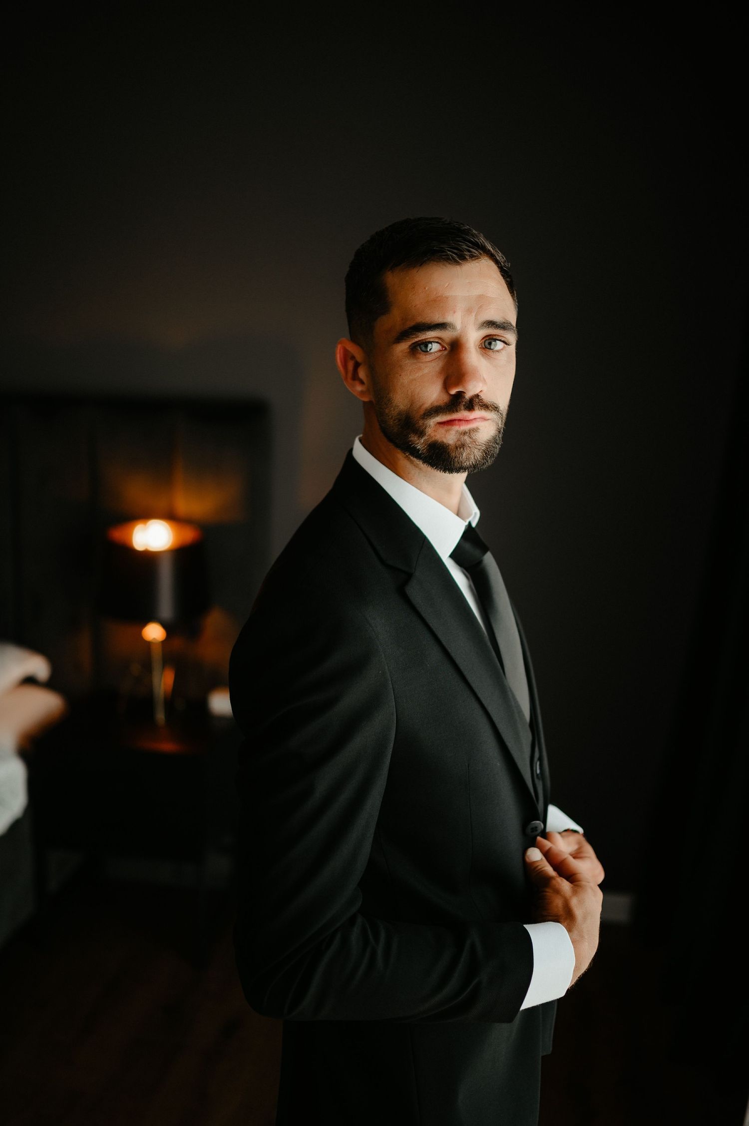 Groom in black tuxedo adjusting jacket in dimly lit room.