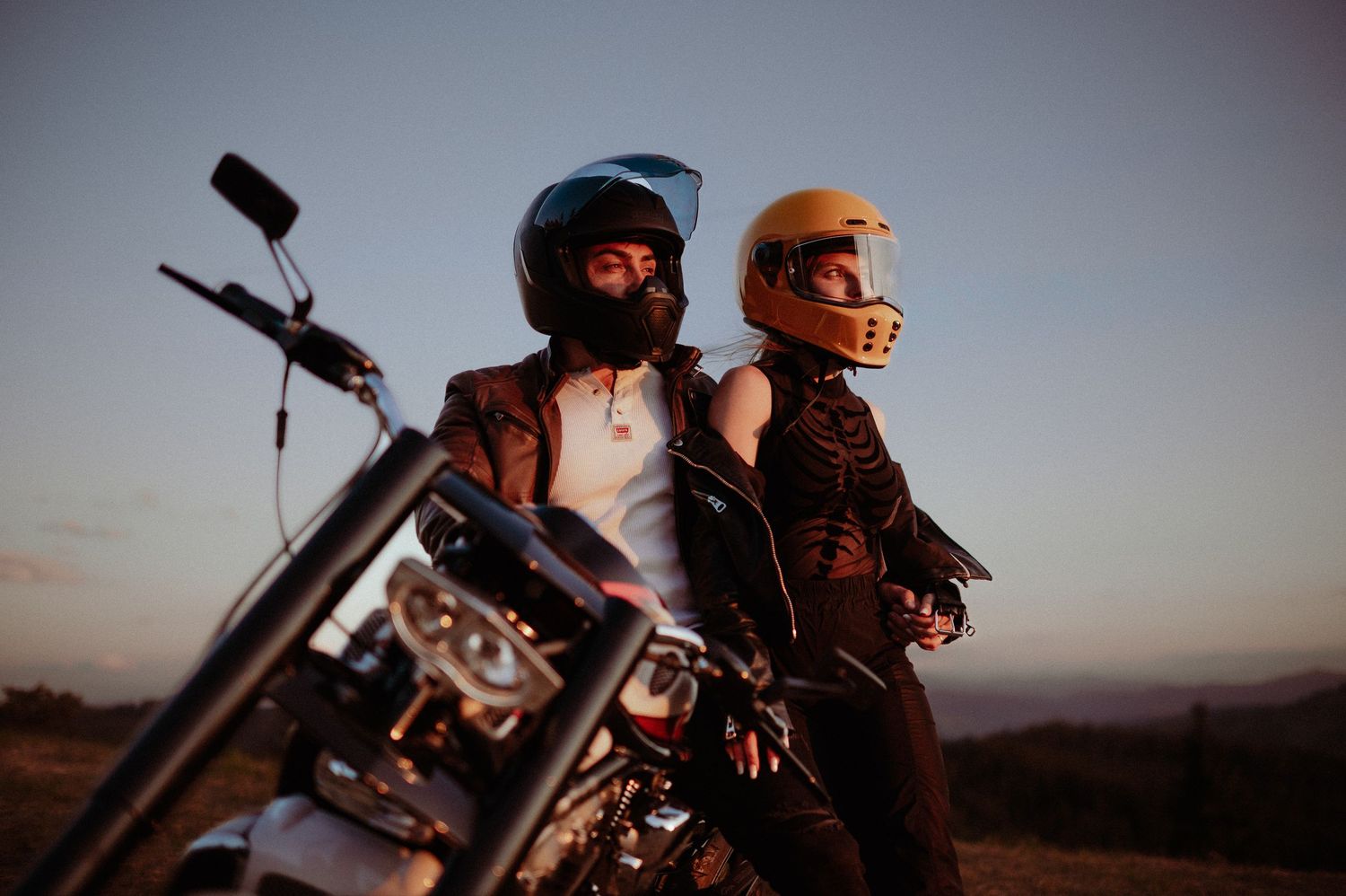 Couple in helmets sitting back-to-back on motorcycle at dusk.