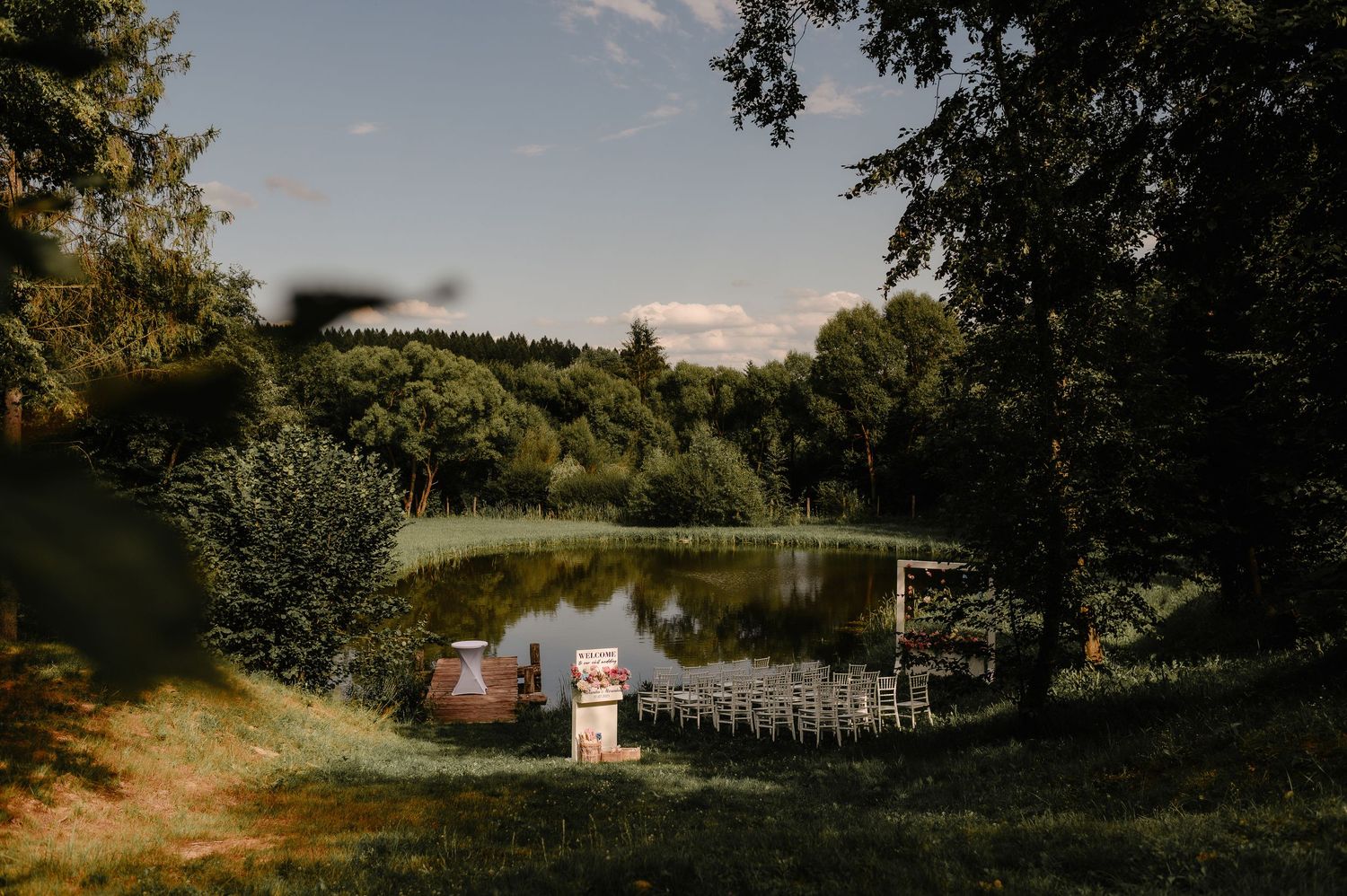 Serene outdoor wedding venue with white chairs arranged near a tranquil pond surrounded by lush trees.