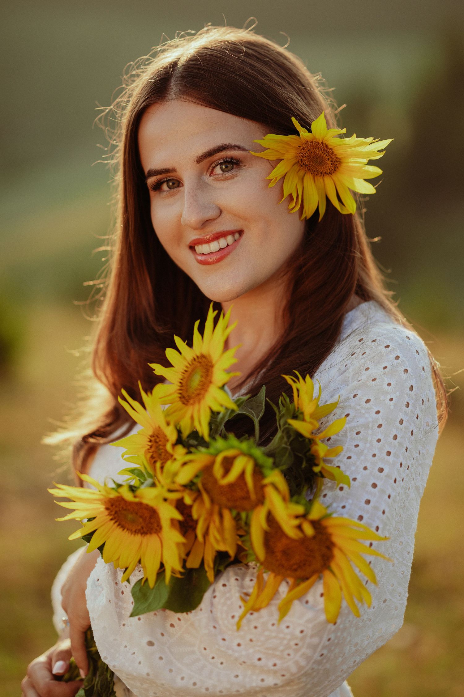 Smiling brunette woman in white dress holding sunflowers with one tucked in her hair.