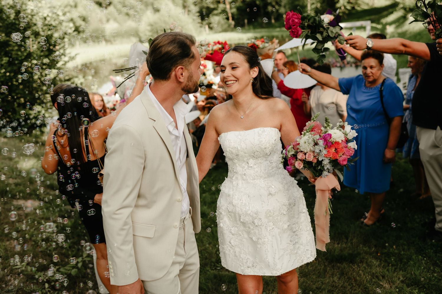 Smiling bride holds colorful bouquet while groom leans in during outdoor wedding celebration.