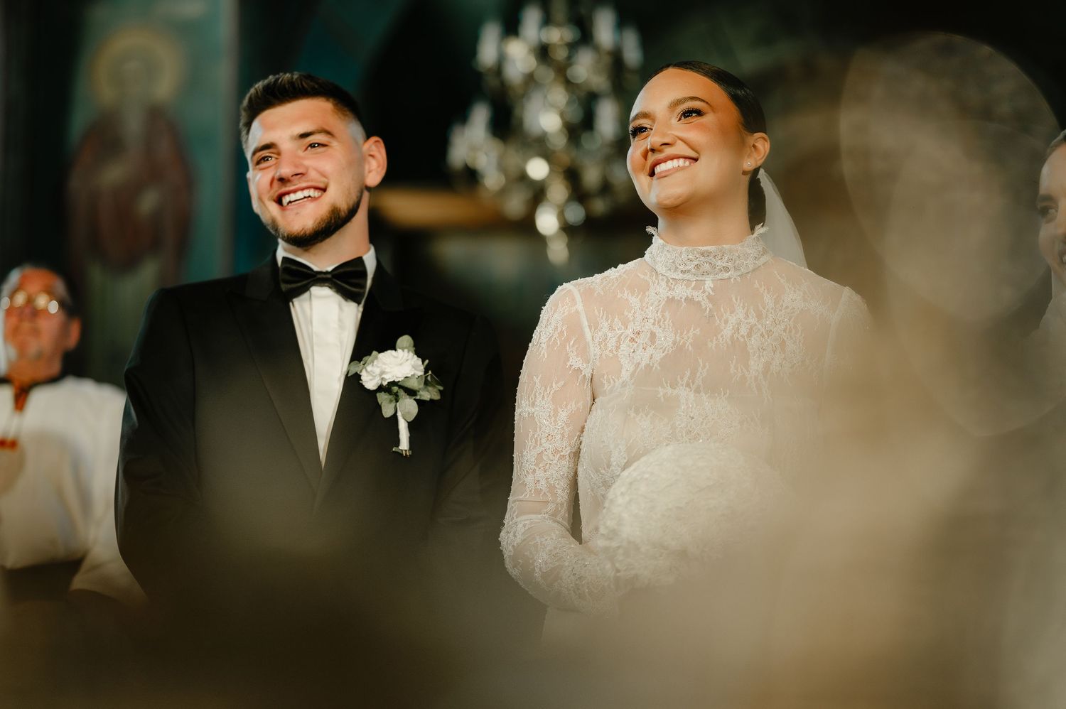 Joyful bride and groom smile during Orthodox wedding ceremony inside beautifully decorated church.