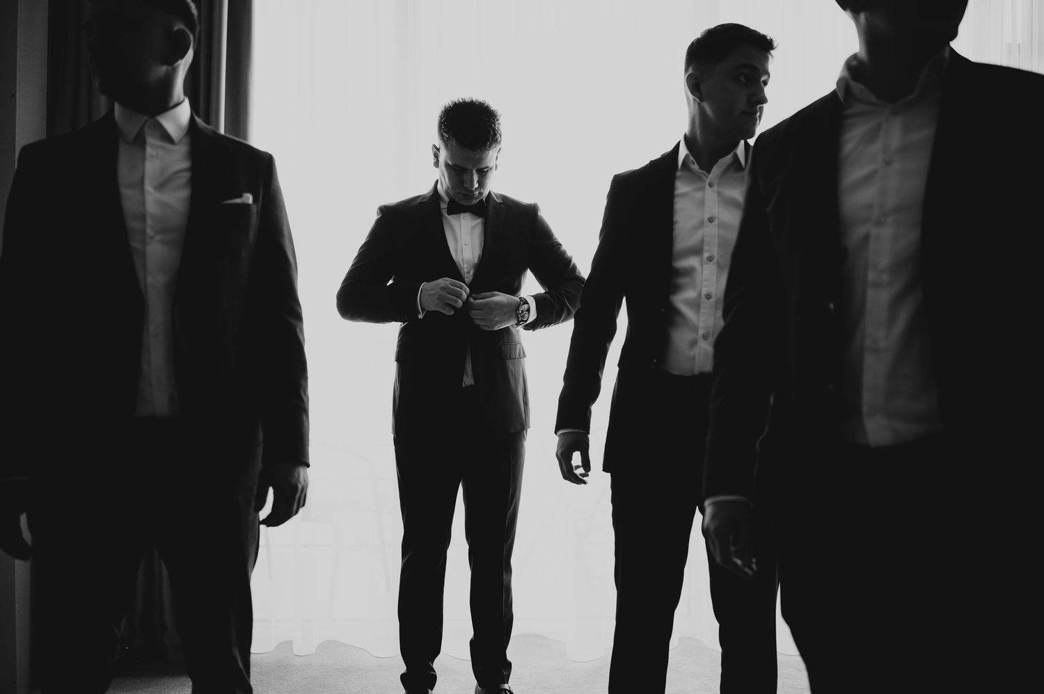 Black and white photo of groom adjusting bow tie flanked by groomsmen in tuxedos.
