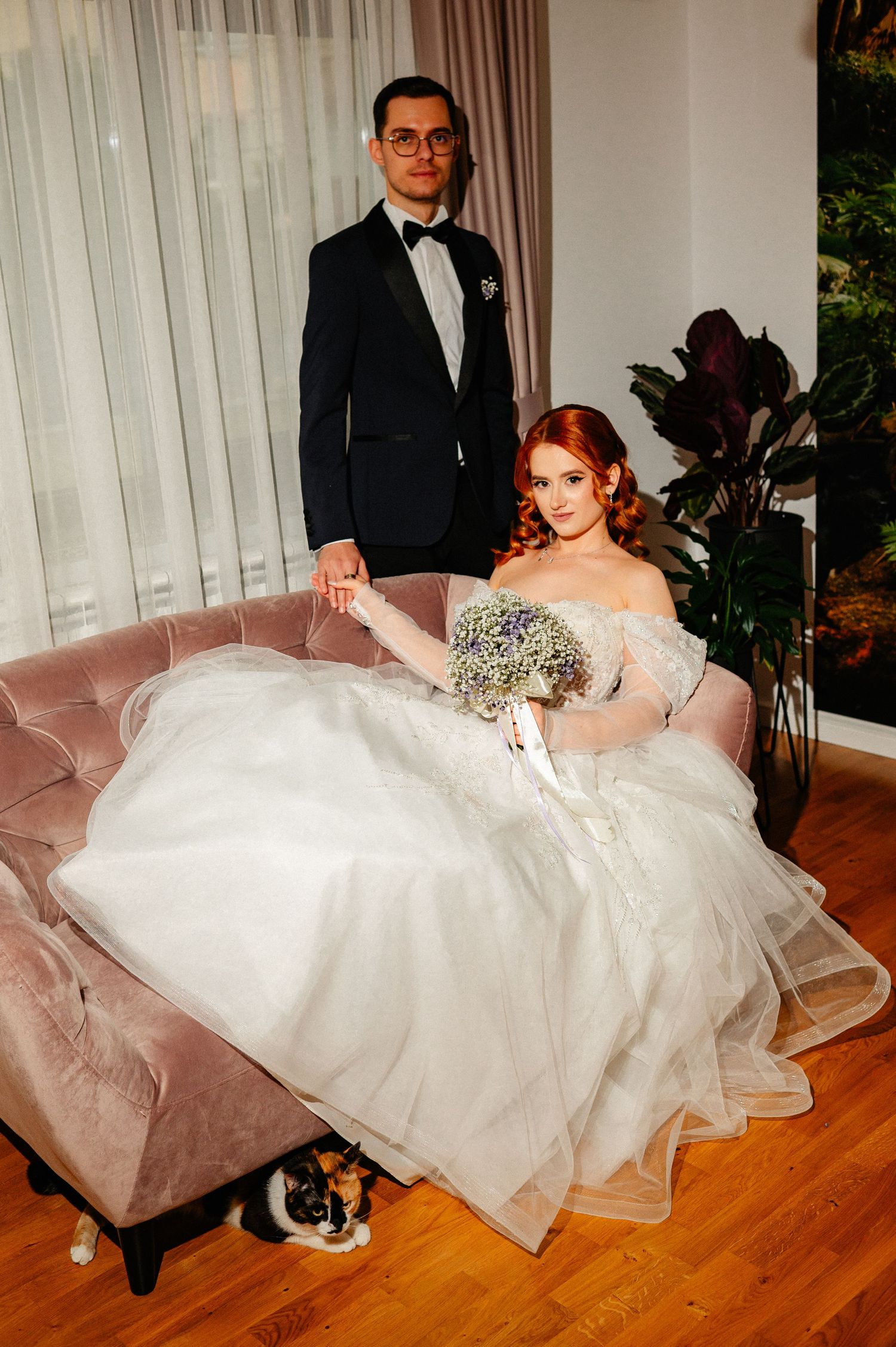 Redhead bride in off-shoulder gown holds bouquet on pink sofa while groom in tuxedo stands beside her.