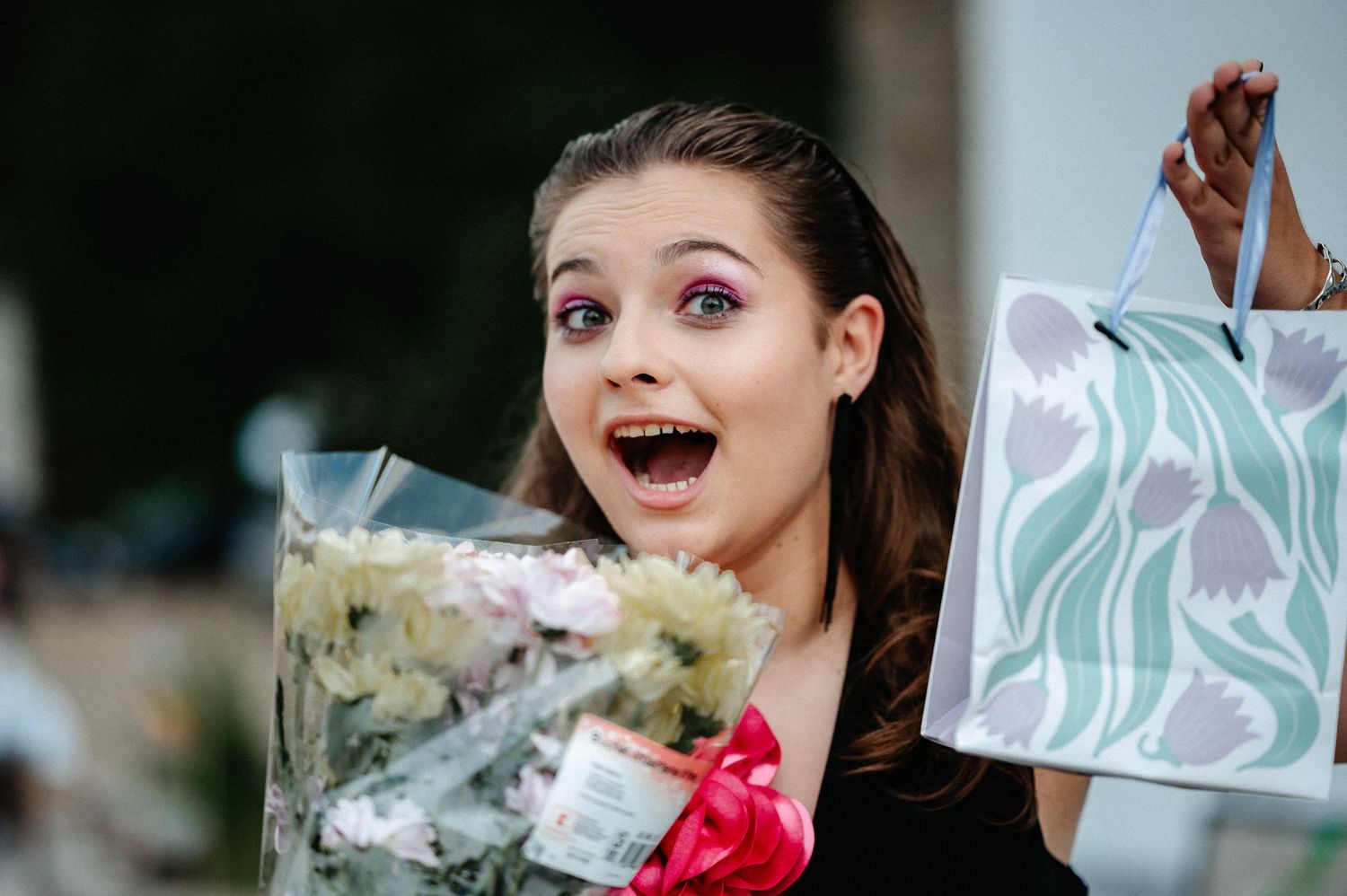 Excited young woman with purple eye makeup holding flowers and a gift bag at an outdoor celebration.
