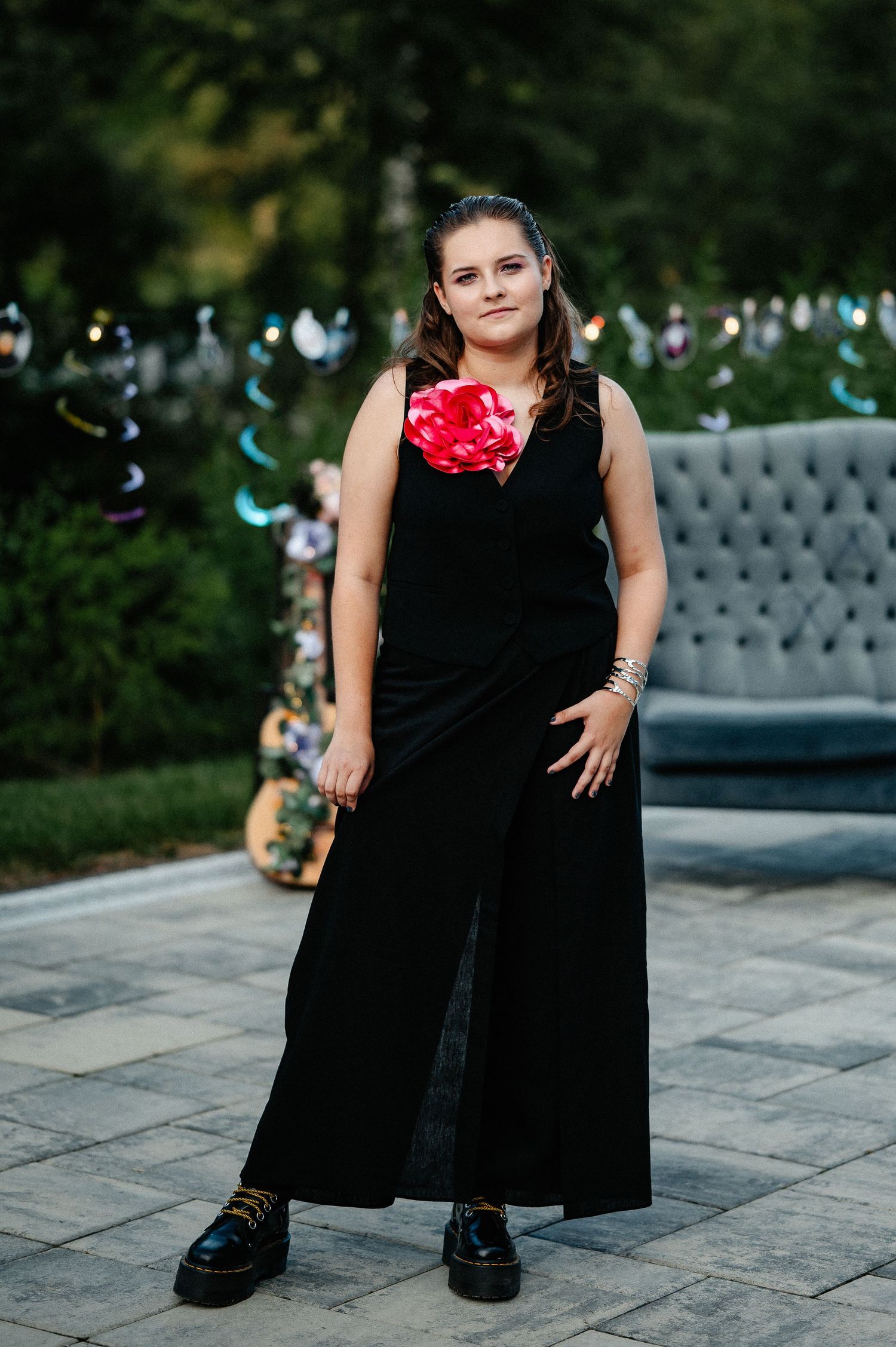 Young woman in black sleeveless maxi dress with red corsage posing outdoors near a gray sofa.