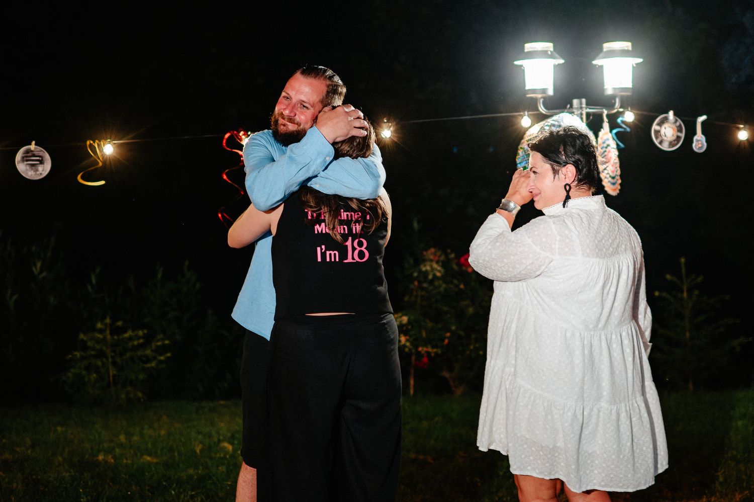A man hugs a girl in an 18th birthday shirt while a woman watches at a night party.