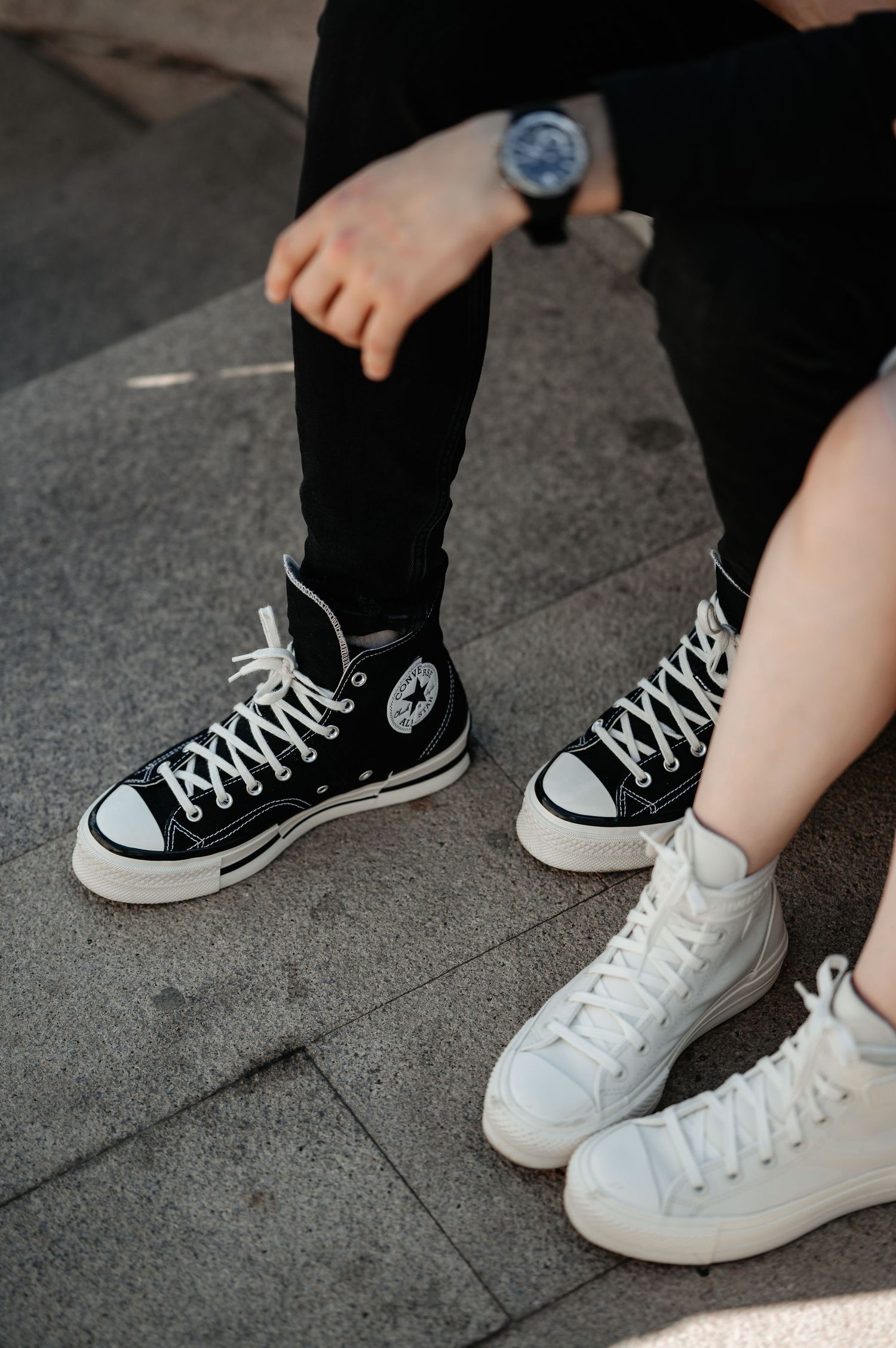 Close-up of two people wearing Converse high-top sneakers, one black and one white, seated on pavement.