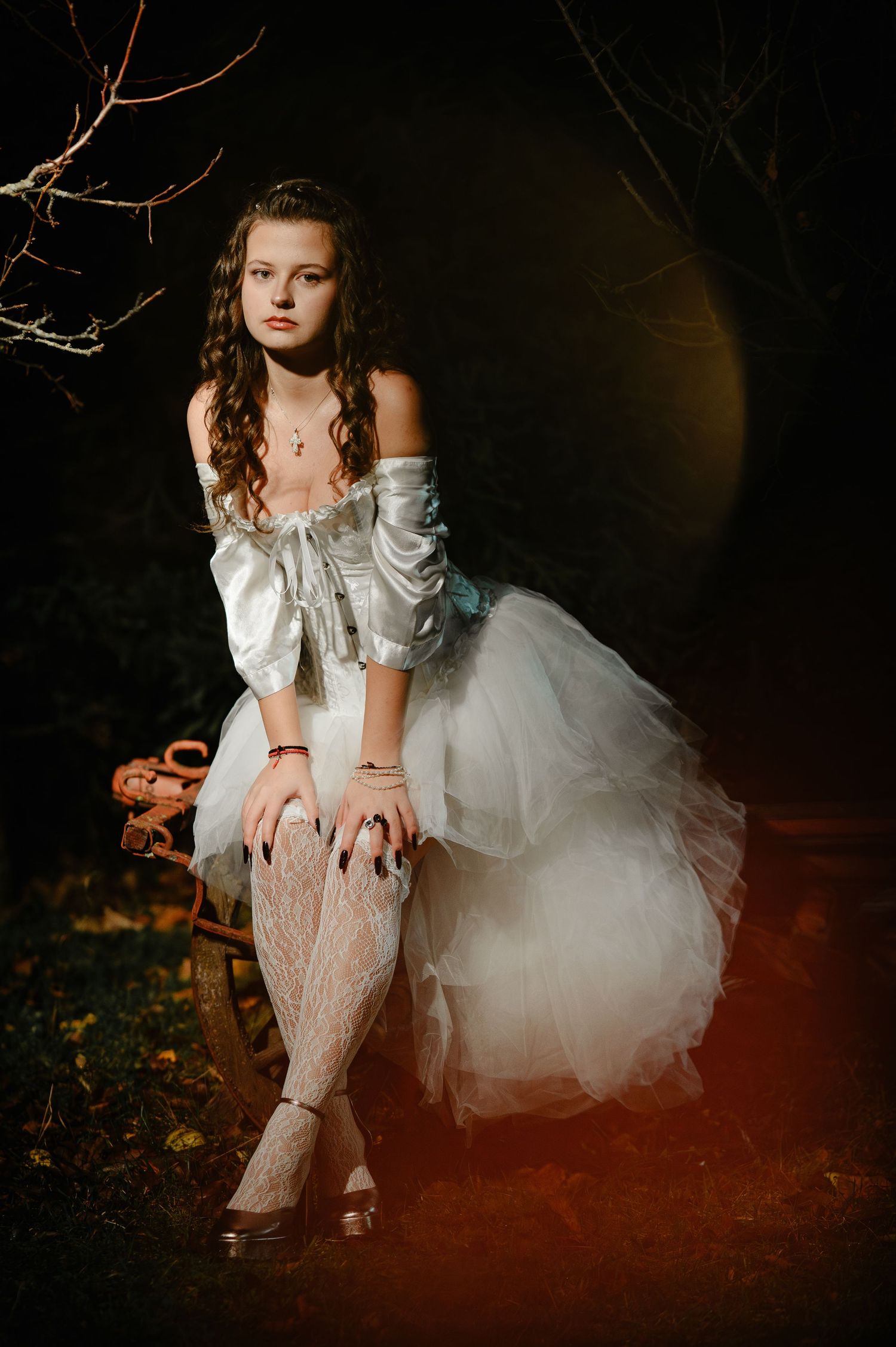 Young woman in white corset gown and lace stockings sits on vintage chair in dark forest setting.