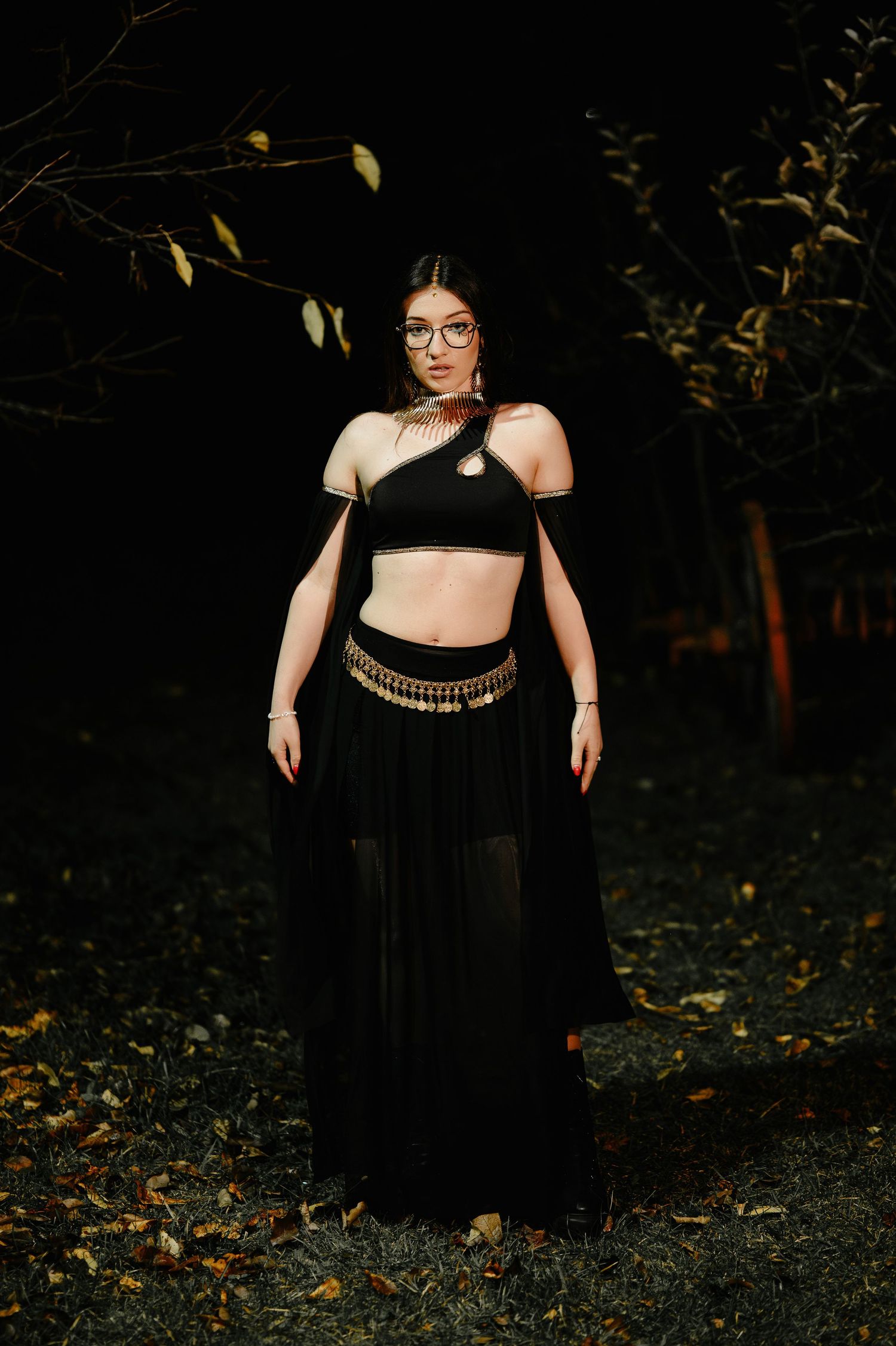 Woman in black belly dance costume with gold belt stands outdoors among autumn leaves.