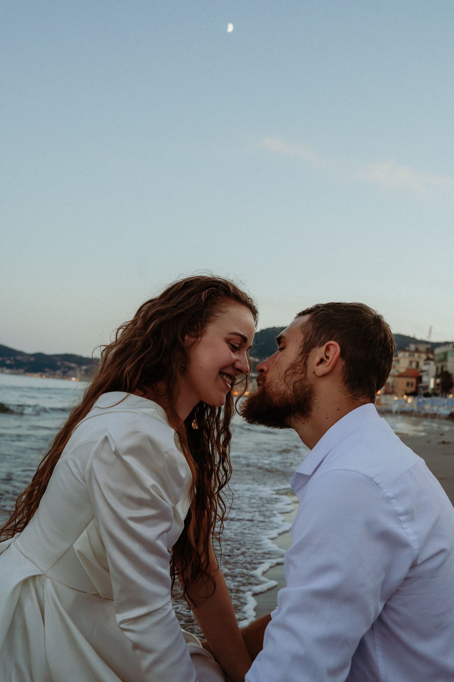 Couple sharing a kiss on a beach at dusk with the moon visible in the sky.