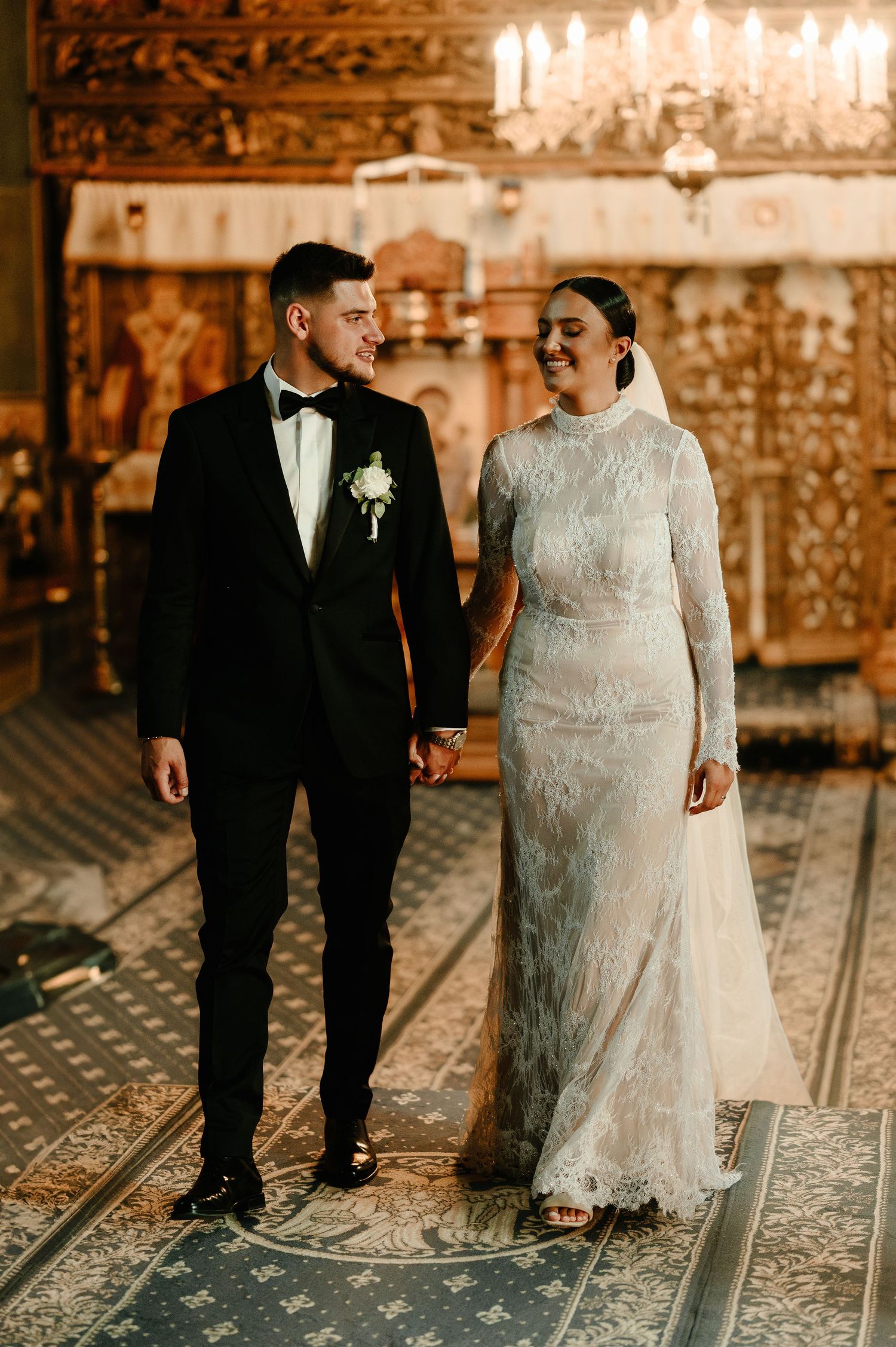 Newlywed couple walking hand in hand smiling at each other inside a grand Orthodox church interior.