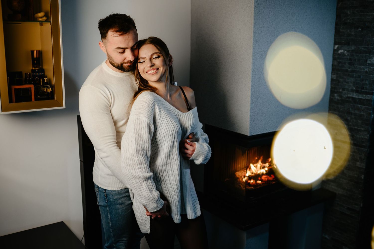 Smiling couple in white sweaters pose together near a fireplace with decorative round lights.