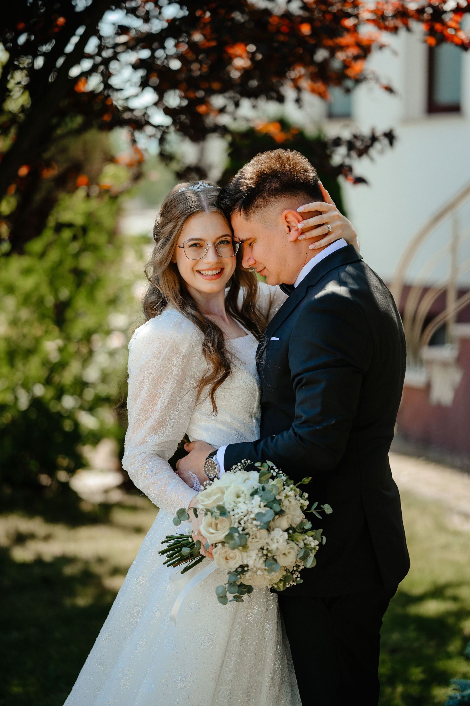 Smiling bride with glasses embracing groom outdoors in golden sunlight with white bouquet.