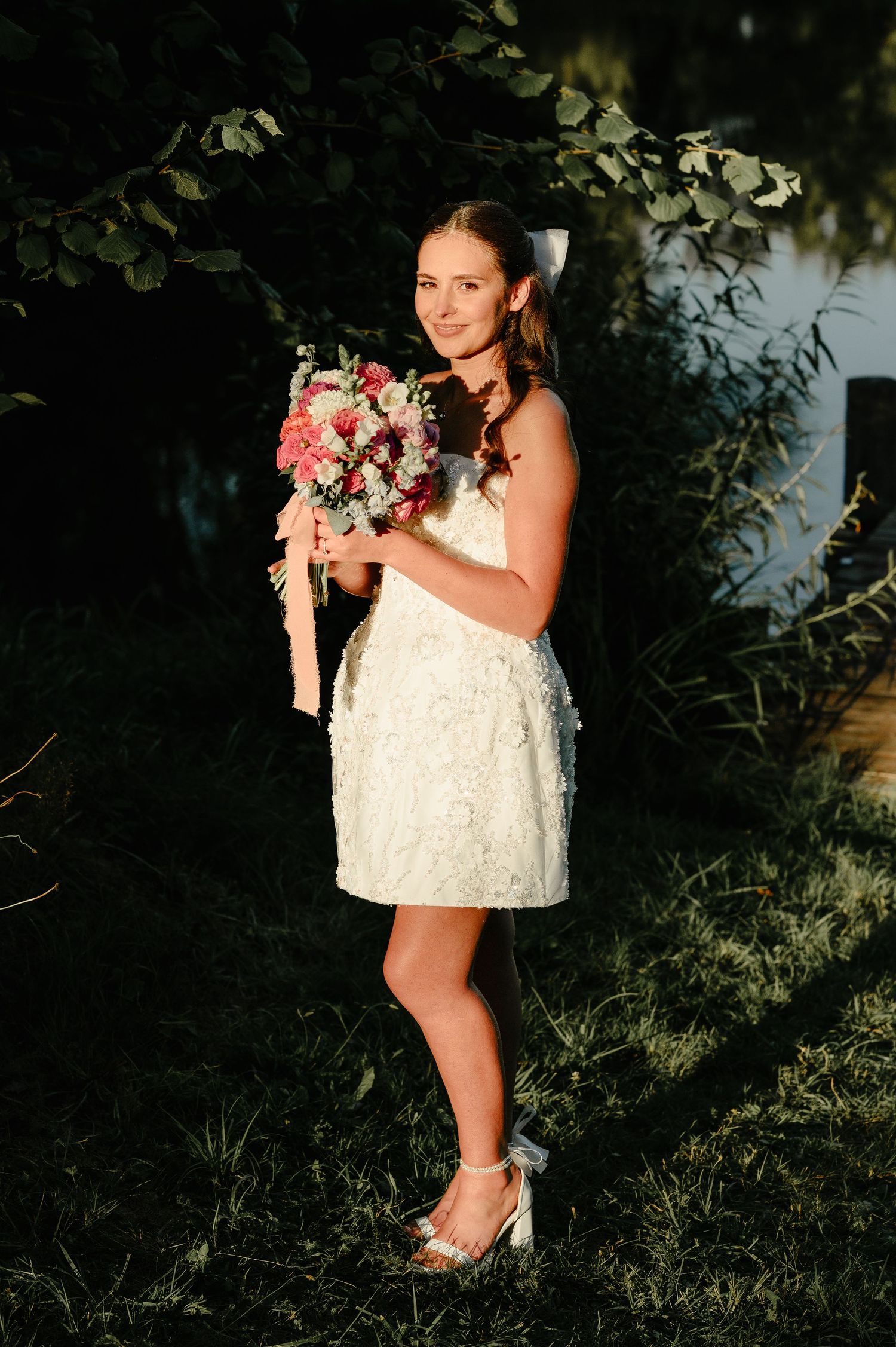 Bride in short white lace dress smiles holding a pink floral bouquet in a sunlit garden setting.