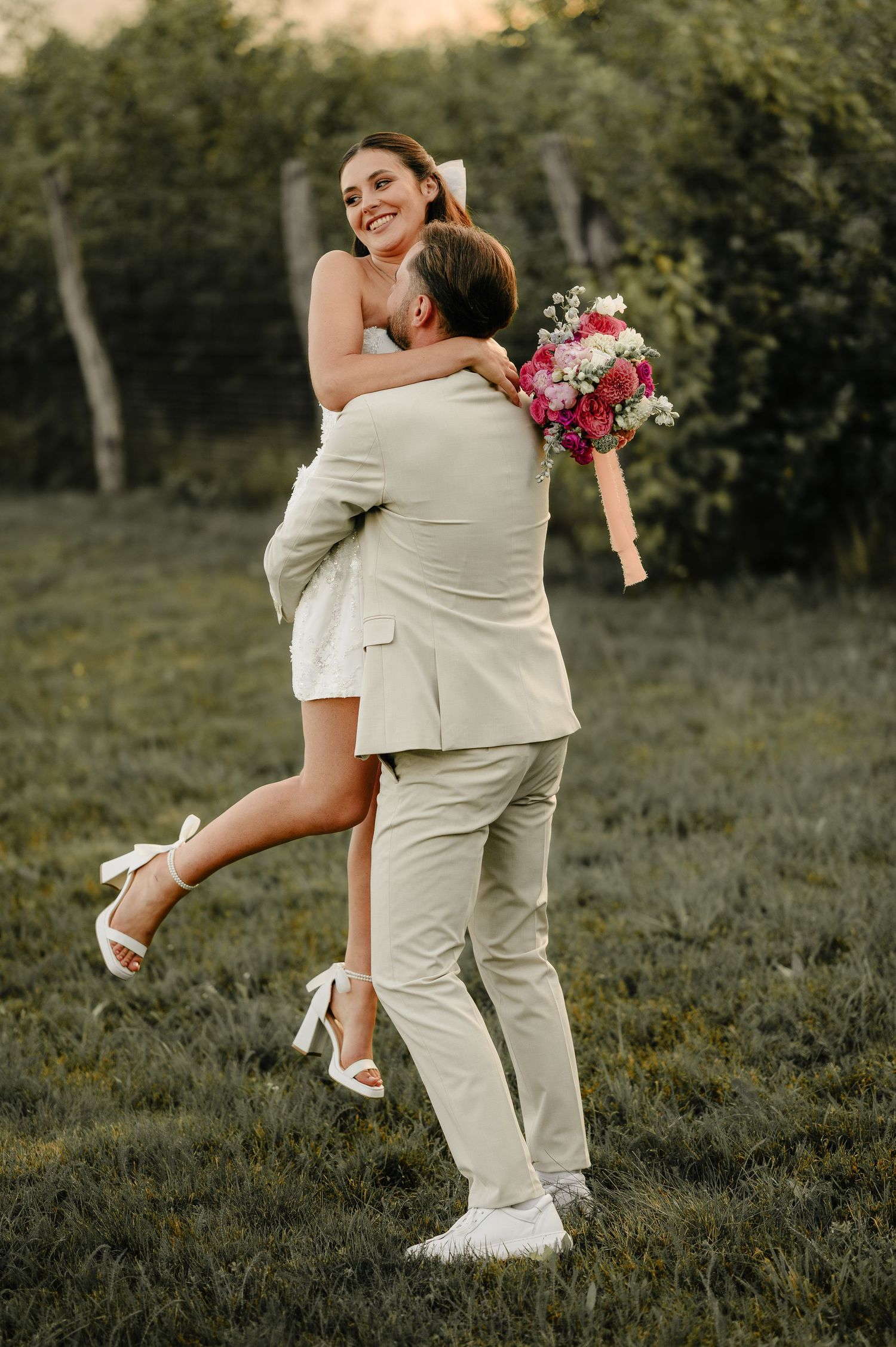 Joyful groom lifting smiling bride in short white dress outdoors in green garden setting.