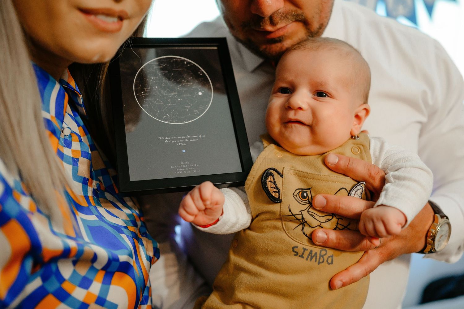 Parents proudly hold smiling baby alongside framed star map print at christening celebration.
