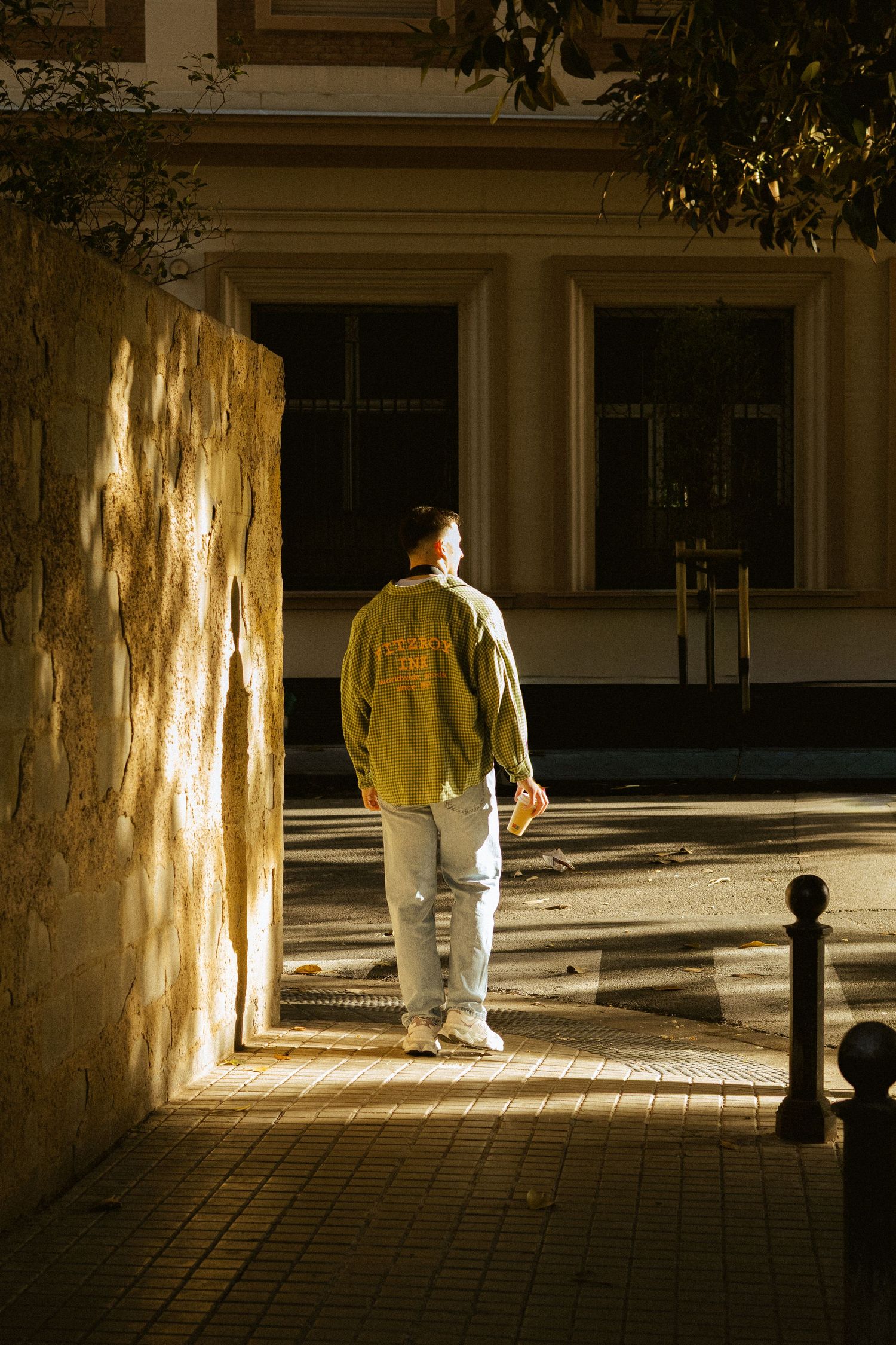 Man walking through a sunlit corridor beside a stone wall near a historic building.