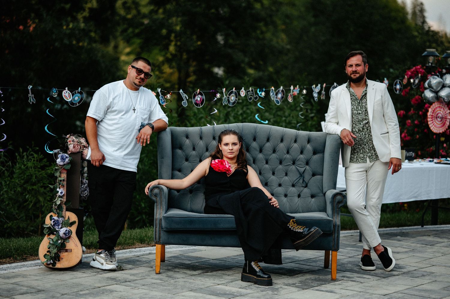 Woman in red dress seated on gray sofa flanked by two men at outdoor garden party.