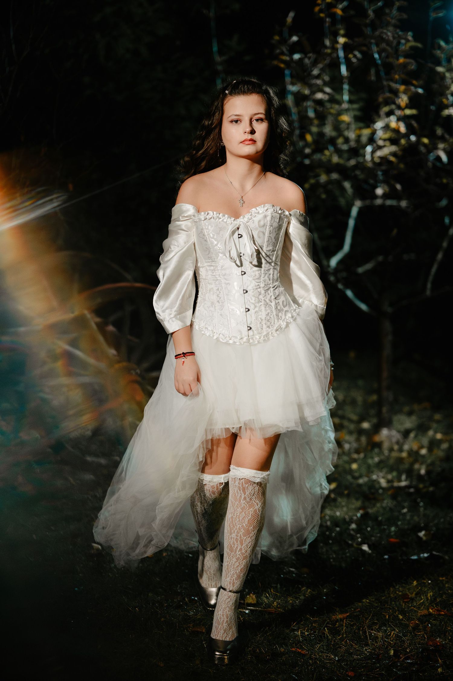 Woman in white off-shoulder corset dress and lace stockings walks through dramatic dark forest scene.