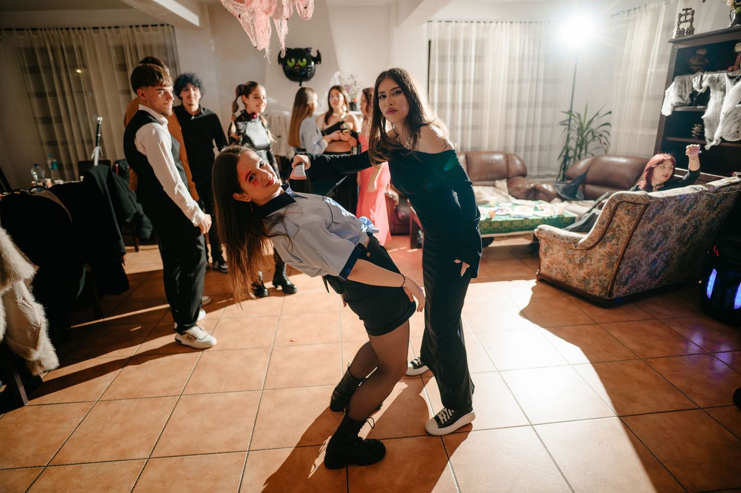 Two young women dance energetically together at an indoor party with guests watching in the background.