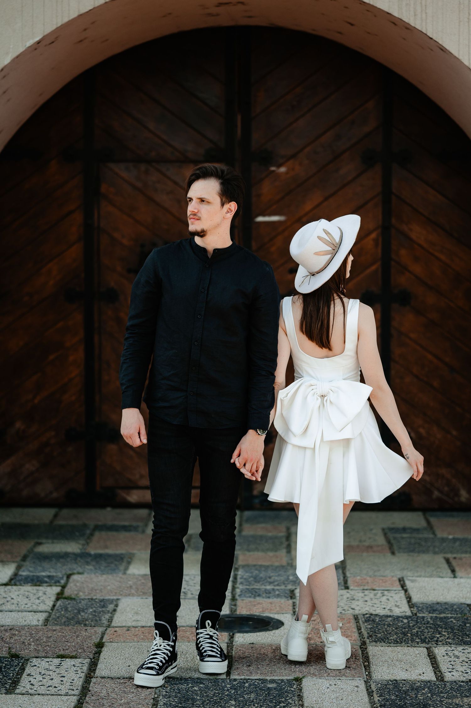Couple holding hands facing a rustic wooden arched door, woman in white backless dress and hat.
