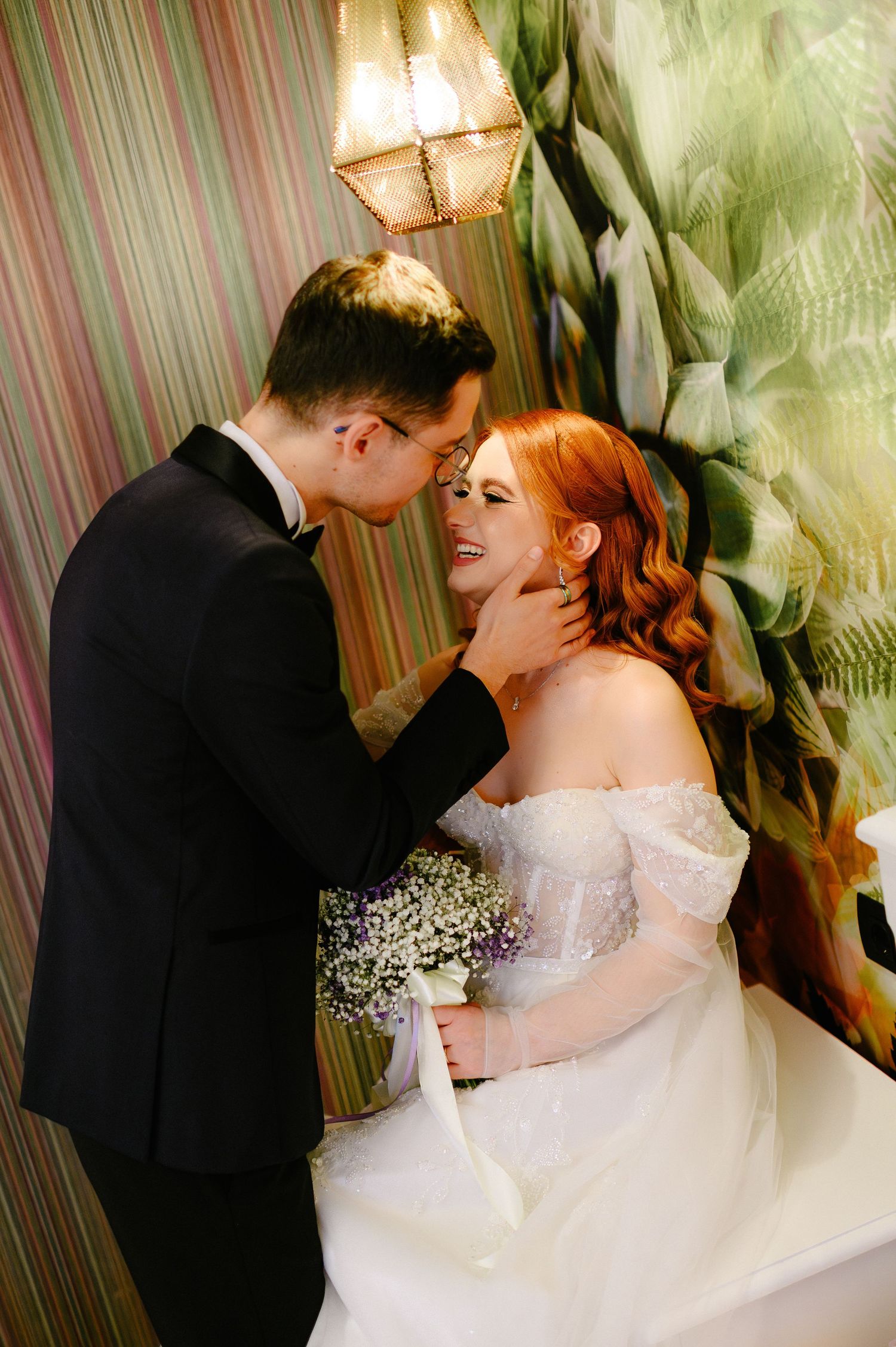 A groom in a black tuxedo tenderly holds a smiling red-haired bride in a white off-shoulder gown holding a bouquet.