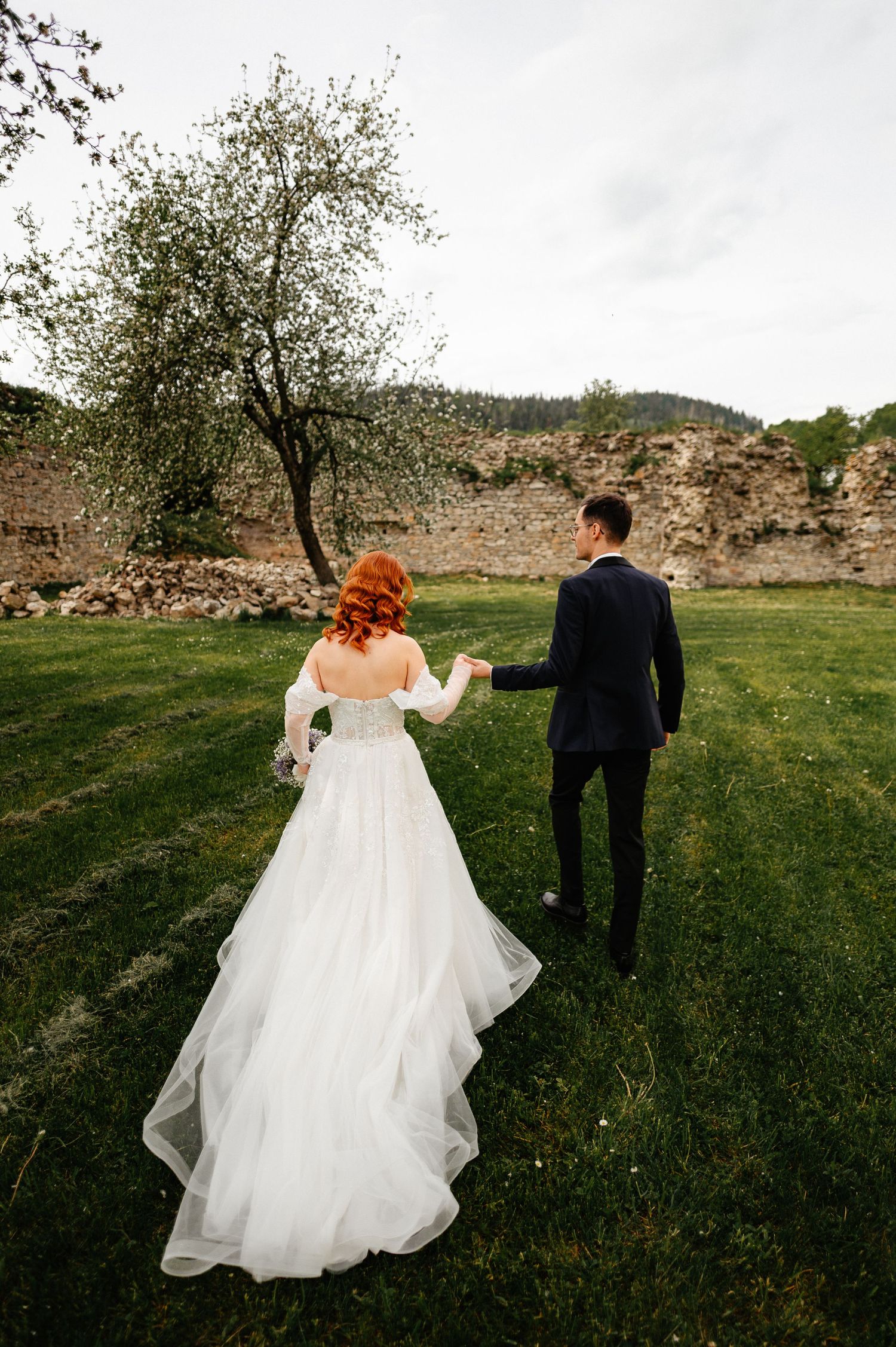 A bride in a white gown with red hair holds hands with a groom in a dark suit walking on green grass.