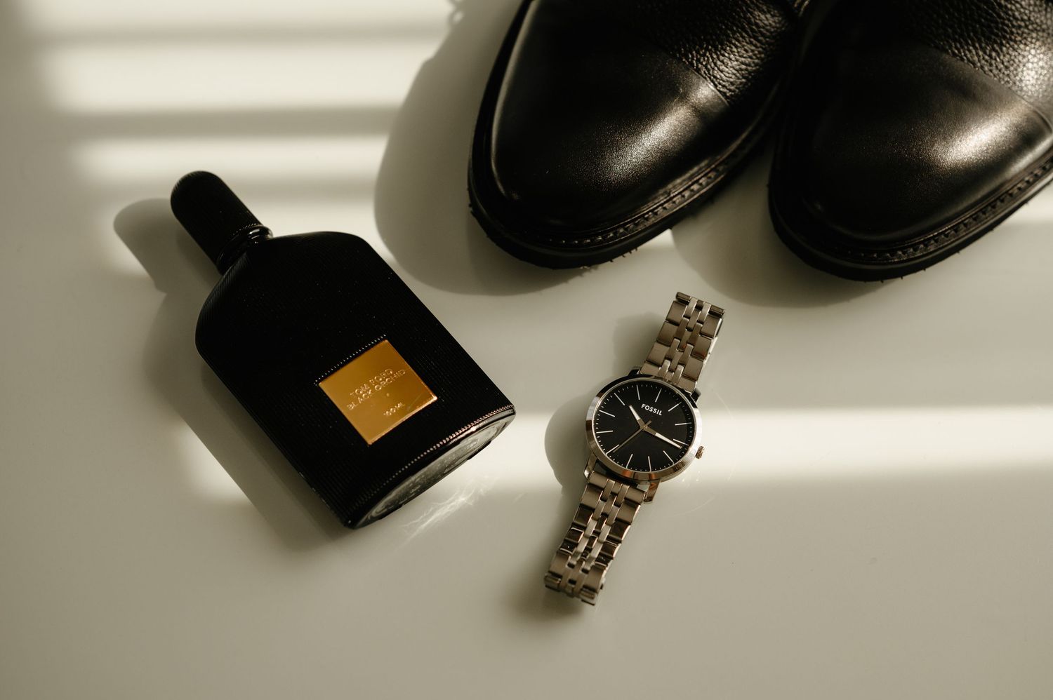 Black leather dress shoes, Tom Ford Black Orchid perfume, and a silver wristwatch arranged on a white surface.