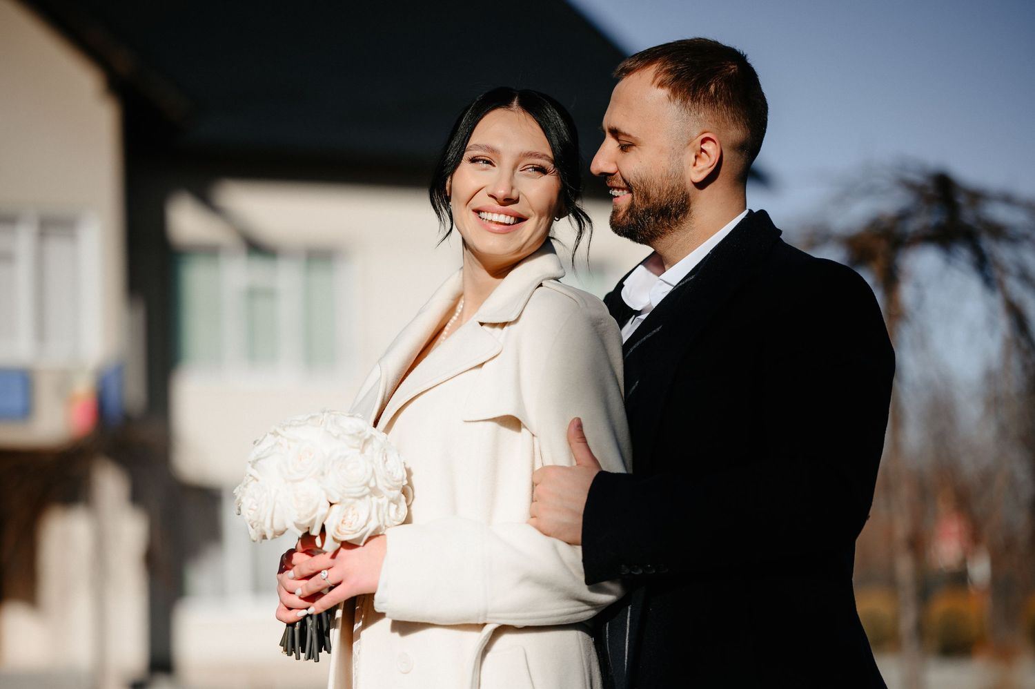 A smiling bride in a white coat holds a bouquet while a groom in a black suit embraces her outdoors on a sunny day.