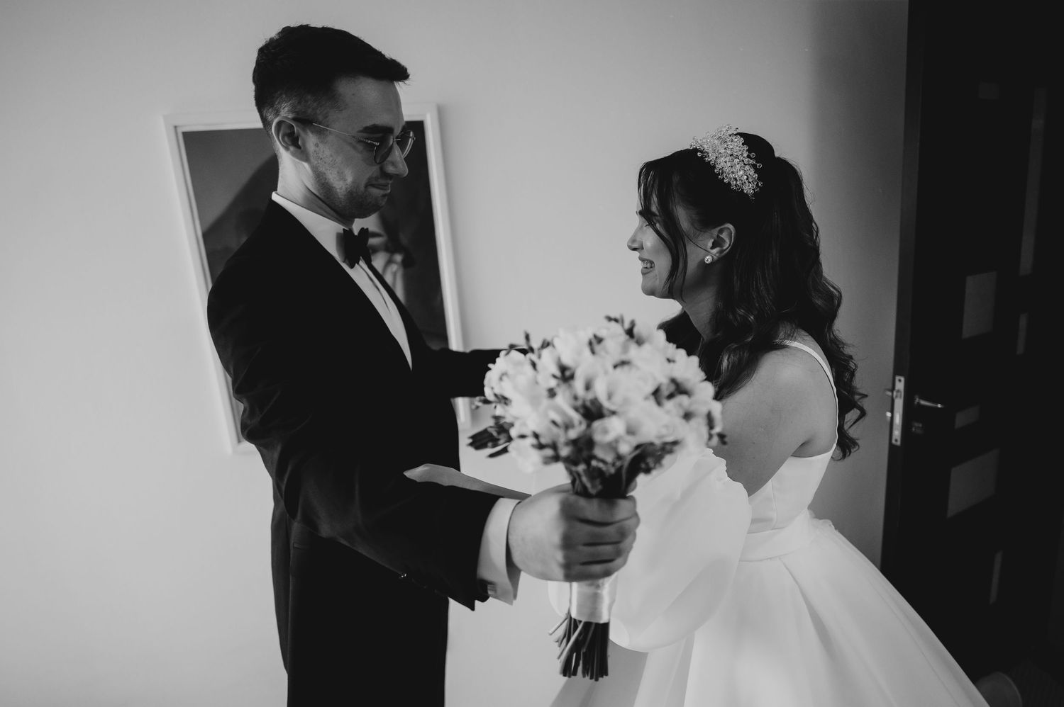 Groom in black tuxedo presents bride with white flower bouquet in black and white wedding photo.