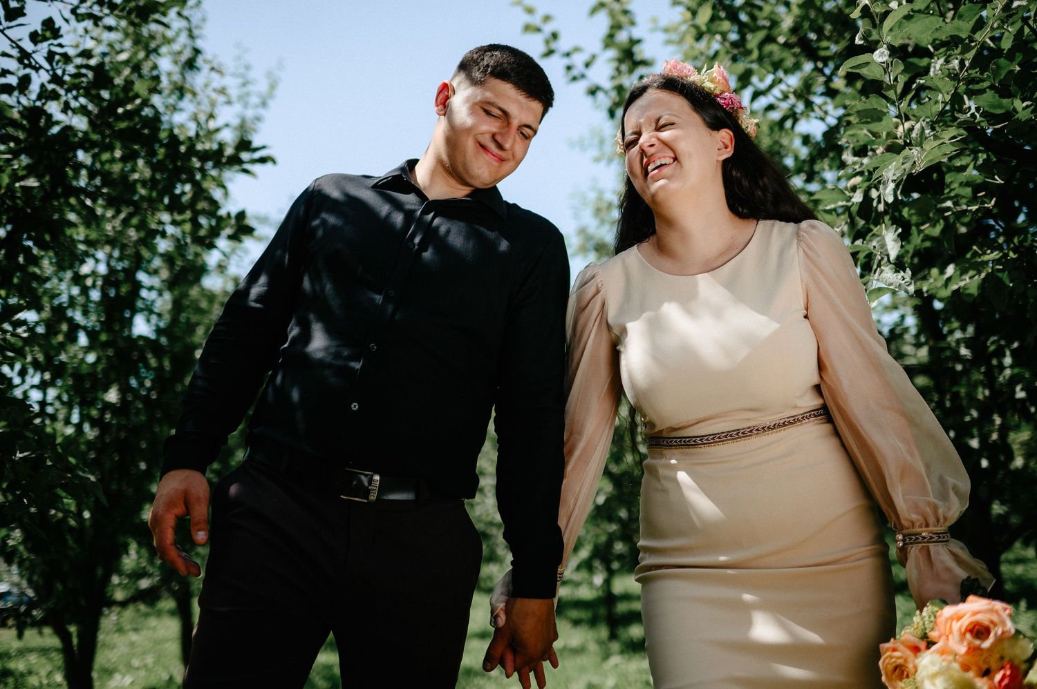 Couple laughing and holding hands in an apple orchard, wearing black leather jacket and champagne wedding dress.