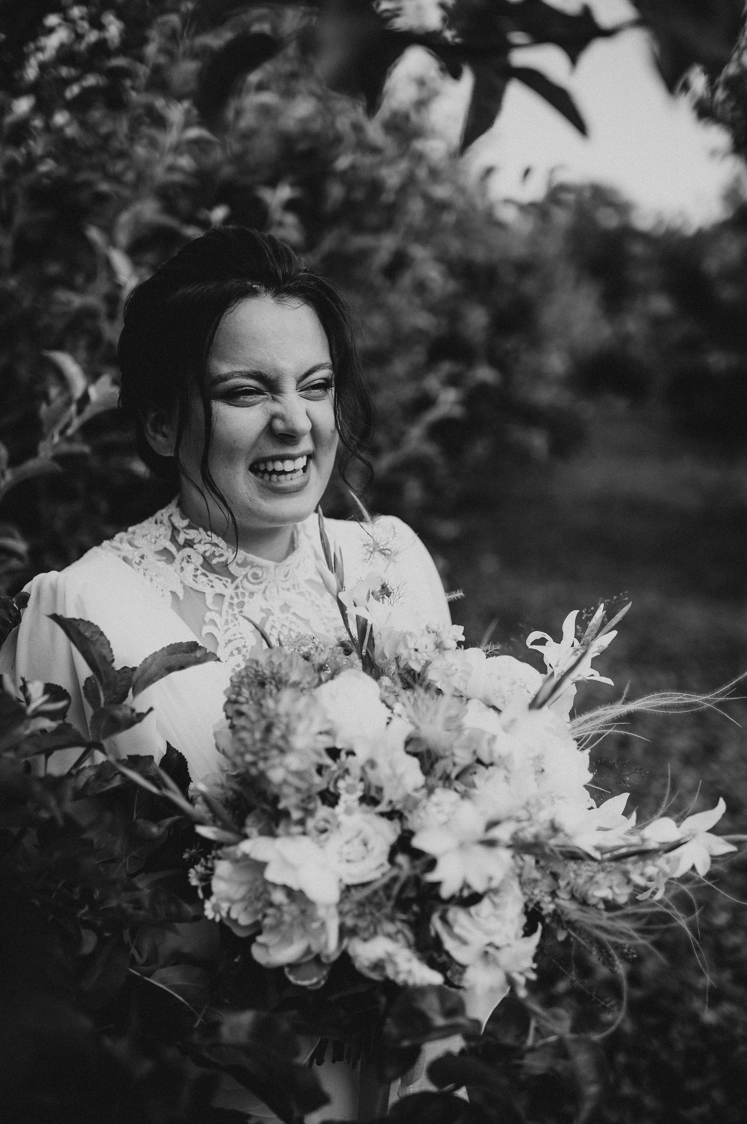 Joyful bride in lace wedding dress laughing while holding a lush floral bouquet in black and white.