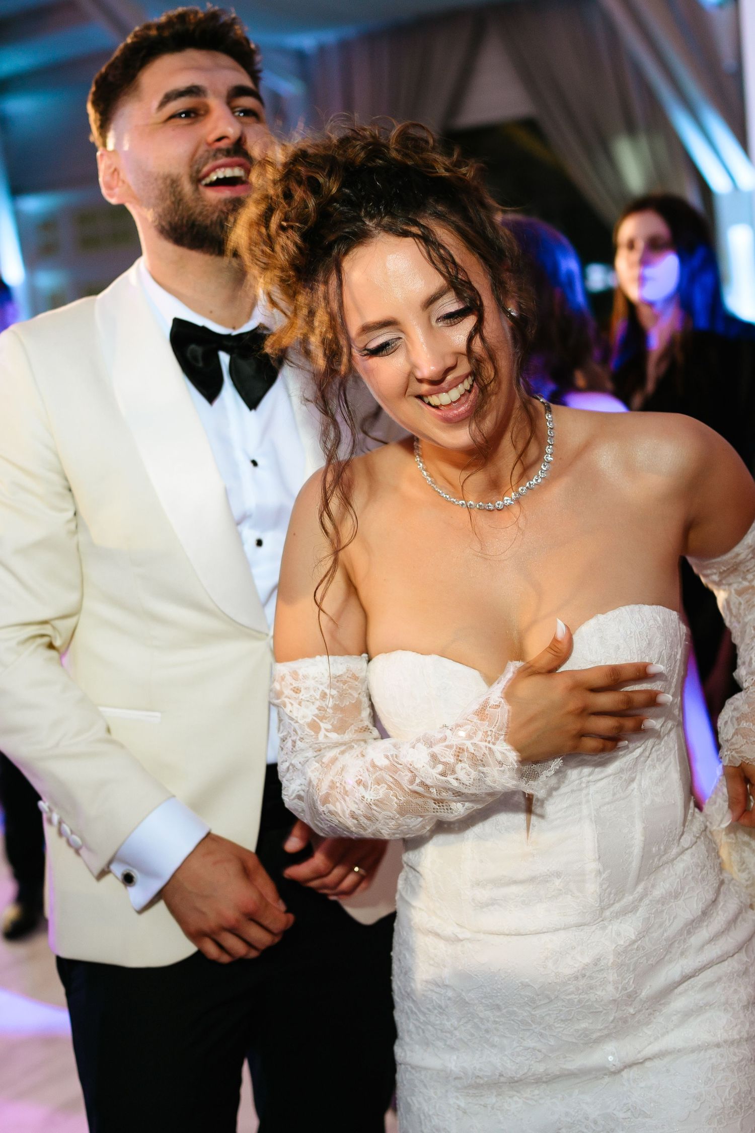 Joyful bride and groom dancing together at wedding reception, wearing white lace gown and ivory tuxedo with bow tie.