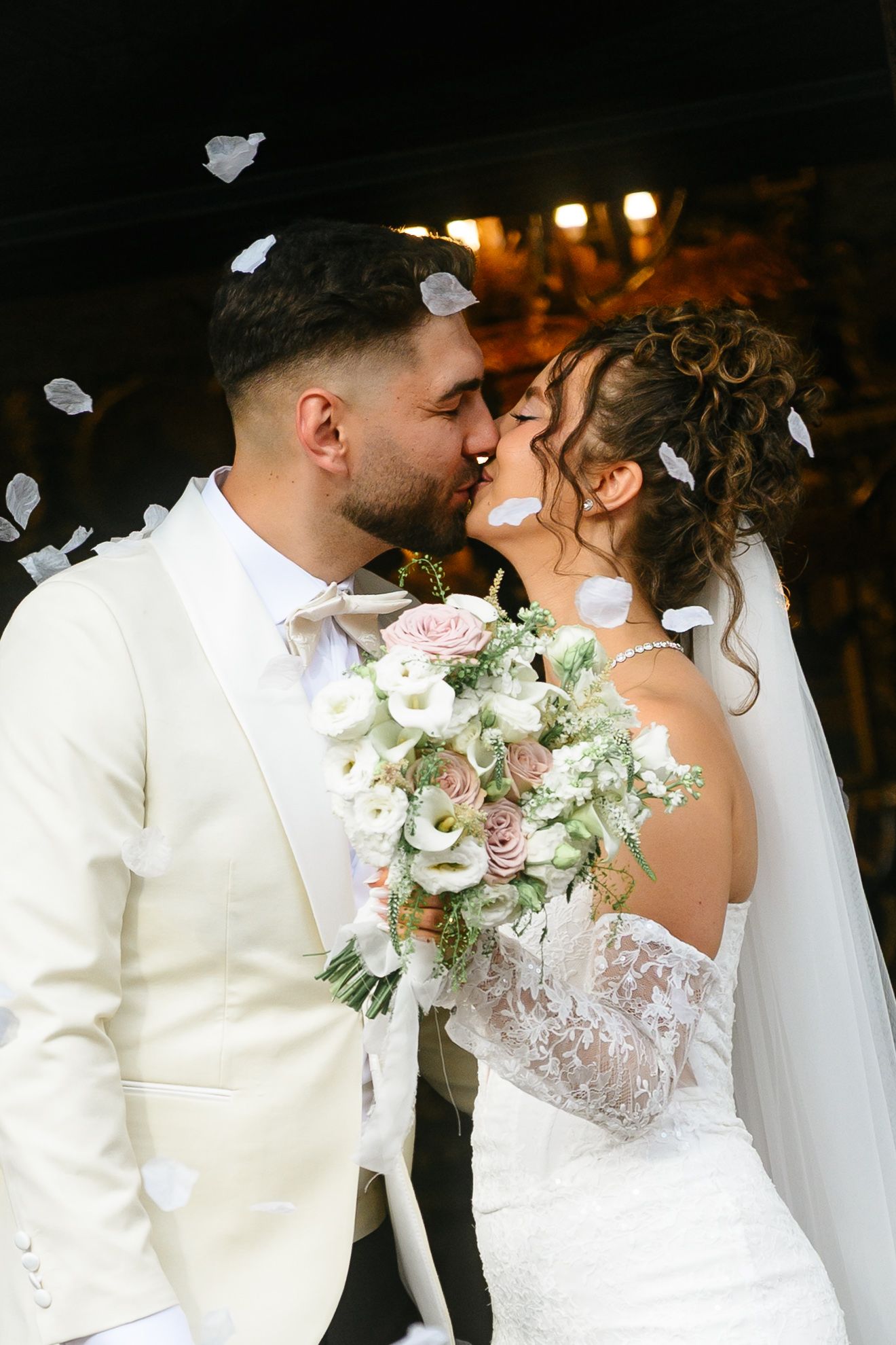 Bride and groom share a romantic kiss with white petals falling around them, bride holding a blush bouquet.