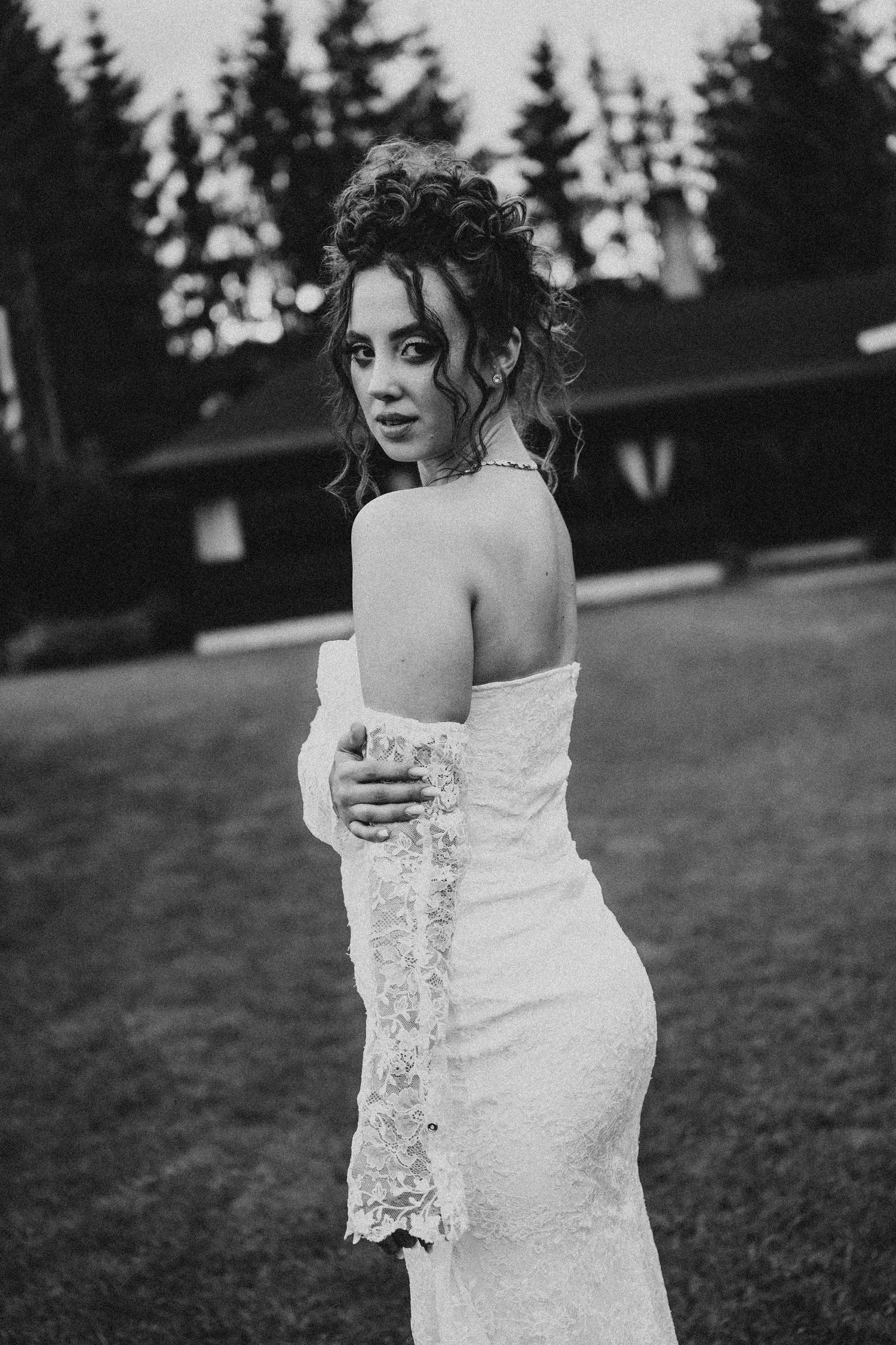 Black and white photo of a bride in a lace wedding gown posing outdoors with trees and a cabin behind her.