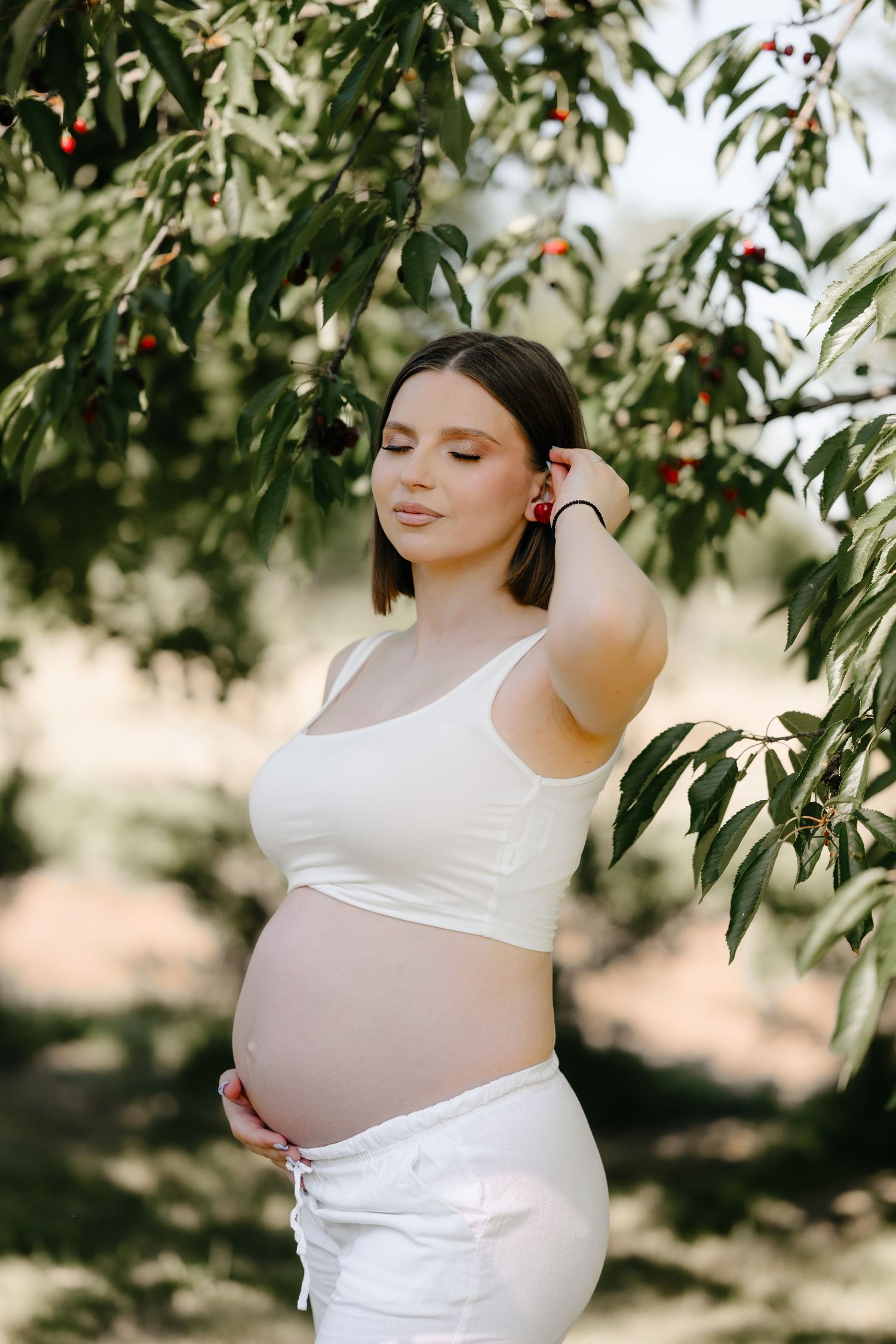 Pregnant woman in white crop top and pants standing under cherry tree, cradling baby bump outdoors.