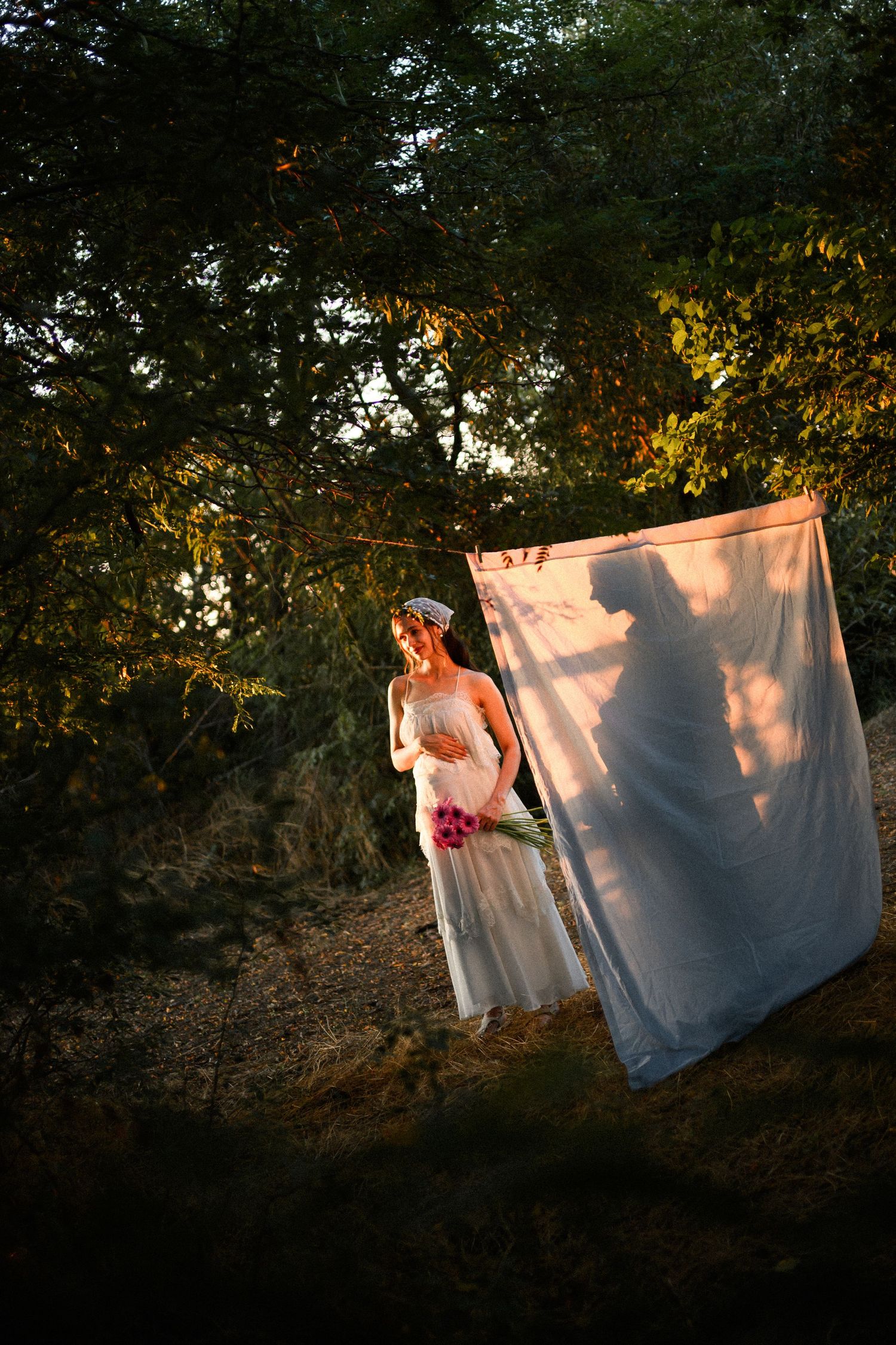 Bride in white gown holding pink flowers stands beside sheer fabric in a sunlit forest setting.