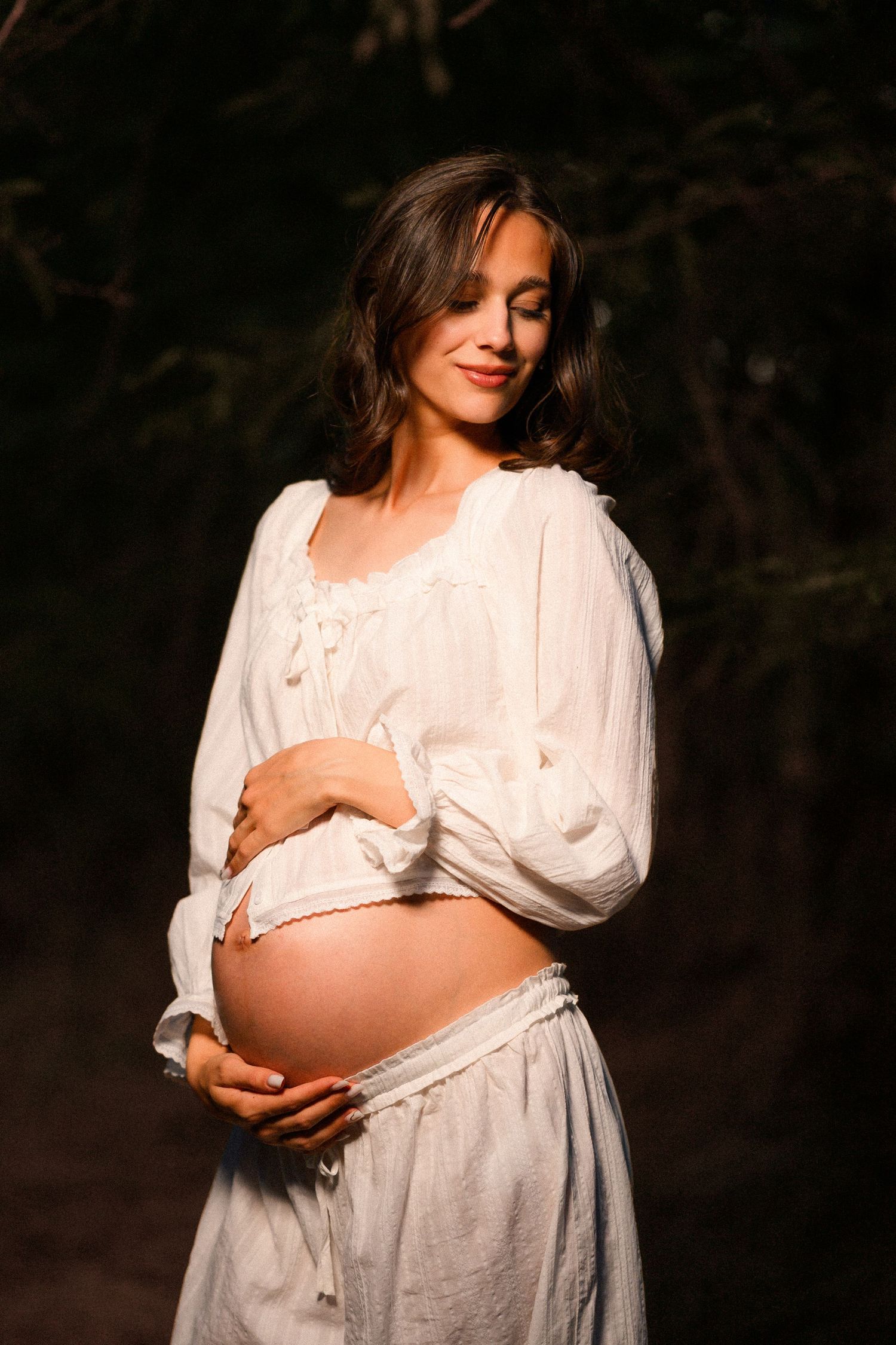 Smiling pregnant woman in white dress cradling her bare baby bump outdoors in a dark forest setting.