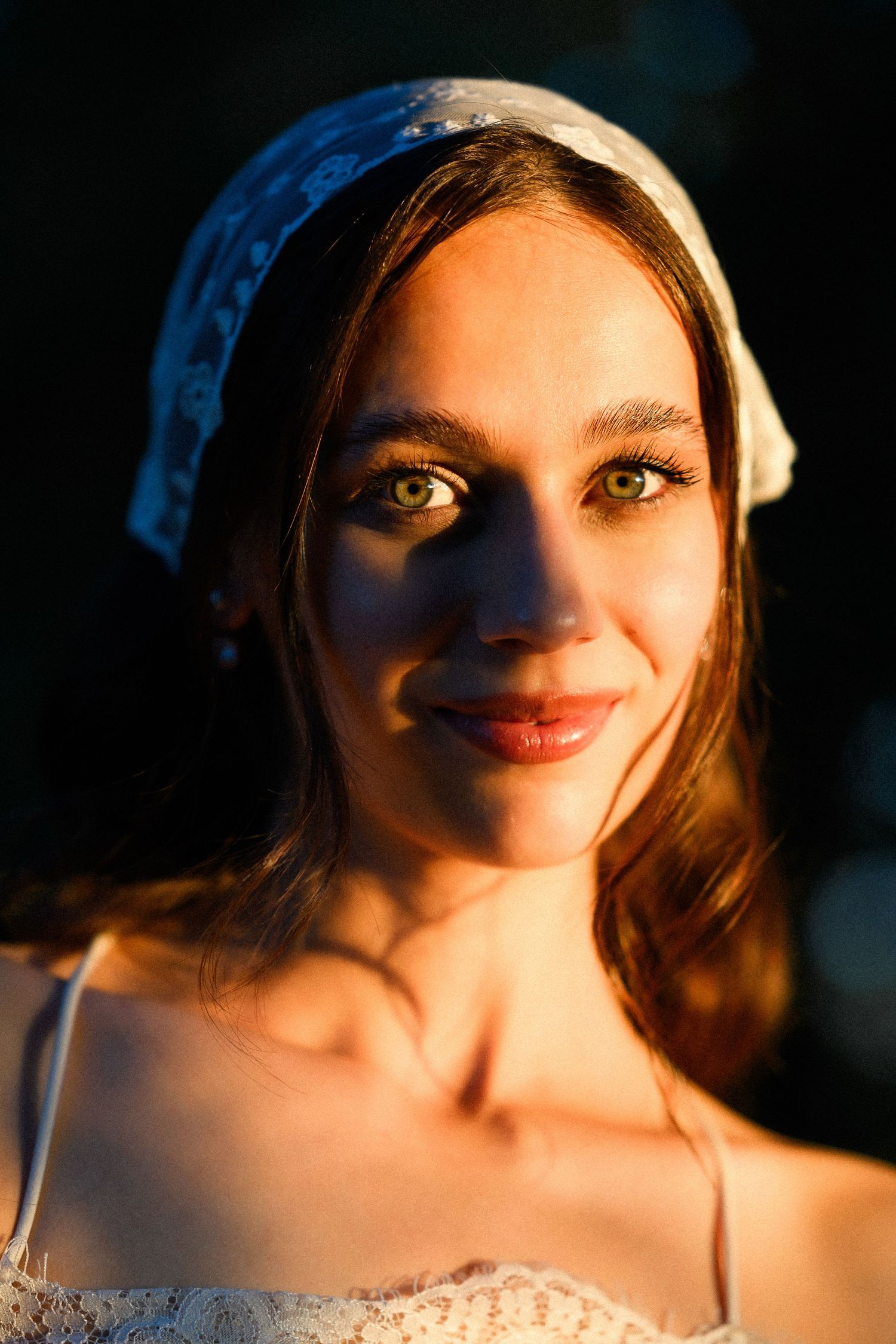 Young woman with green eyes wearing a white headscarf, bathed in warm golden light against a dark background.
