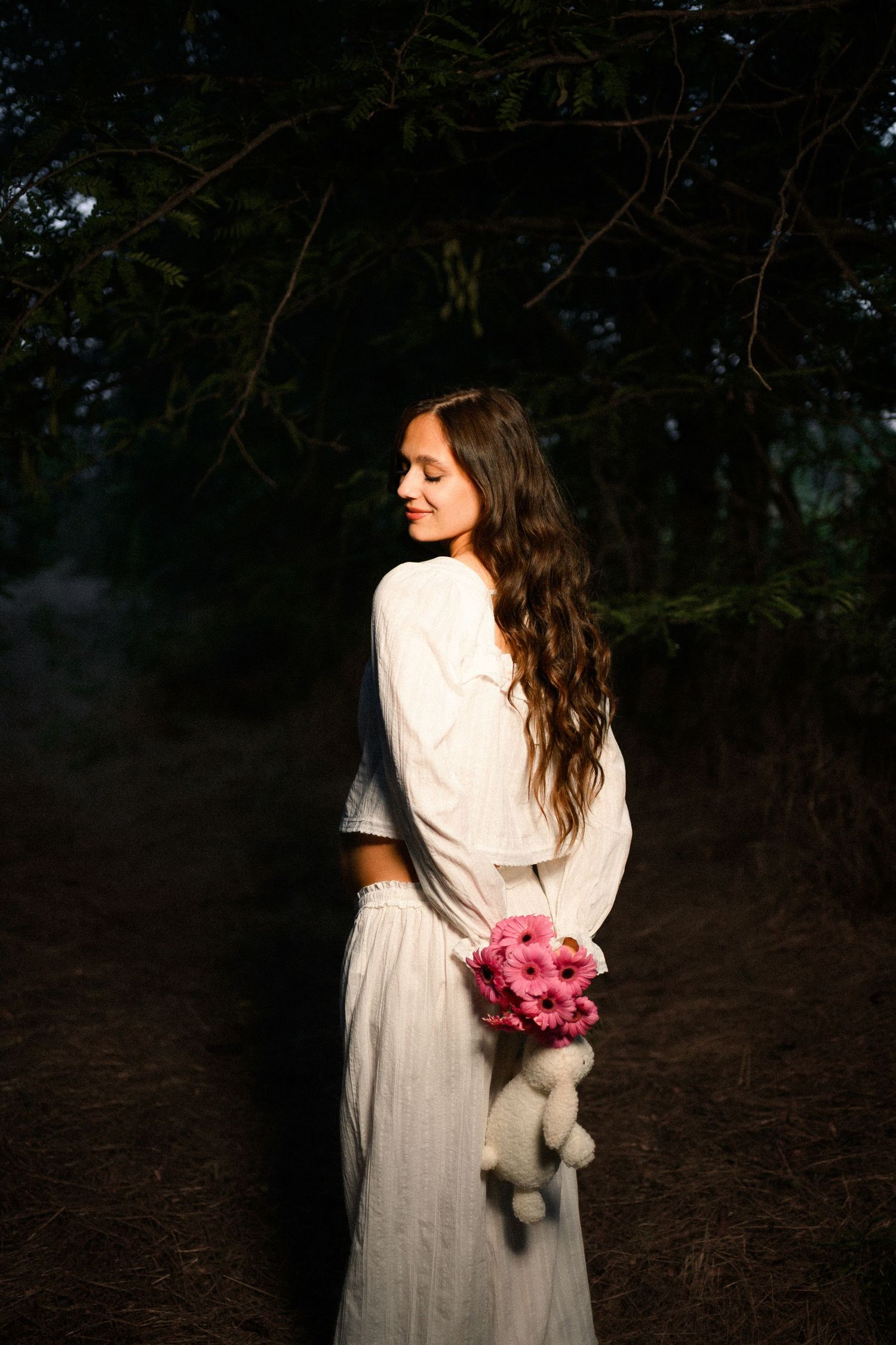 Young woman in white dress holding pink flowers and teddy bear, standing on a dark forest path at dusk.