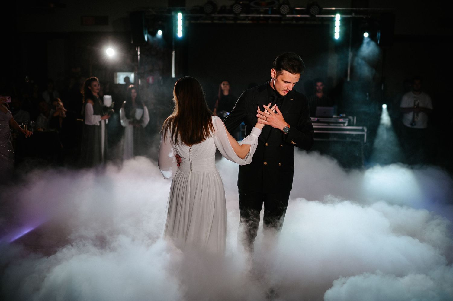 Bride and groom hold hands tenderly during first dance surrounded by thick white fog on dance floor.