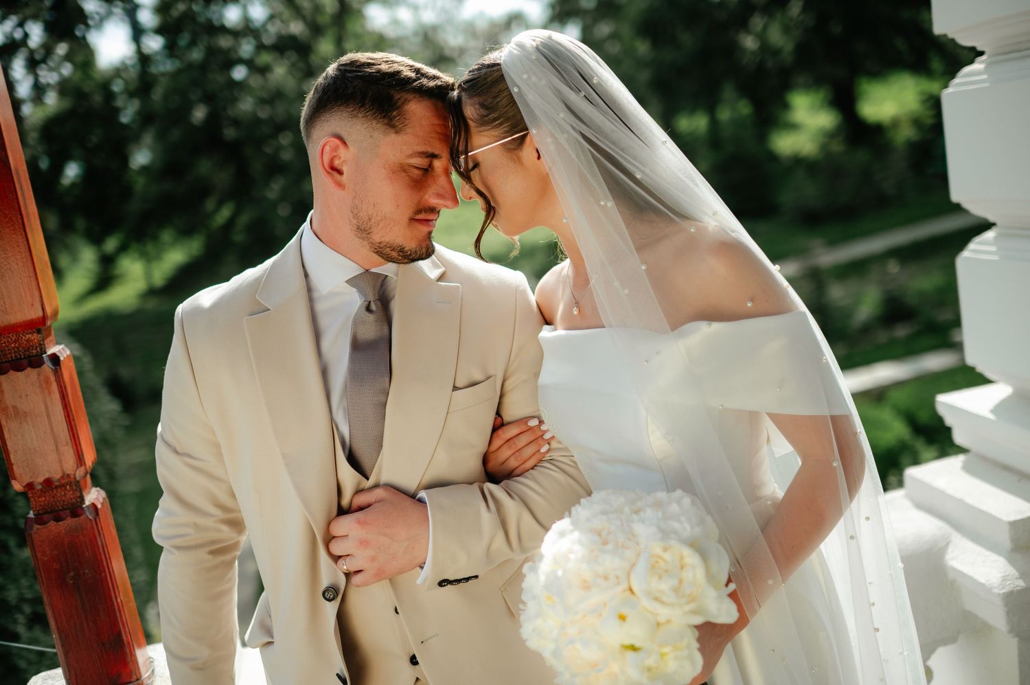 Bride and groom share an intimate moment on balcony, foreheads touching, bride holding white bouquet in beige suit.