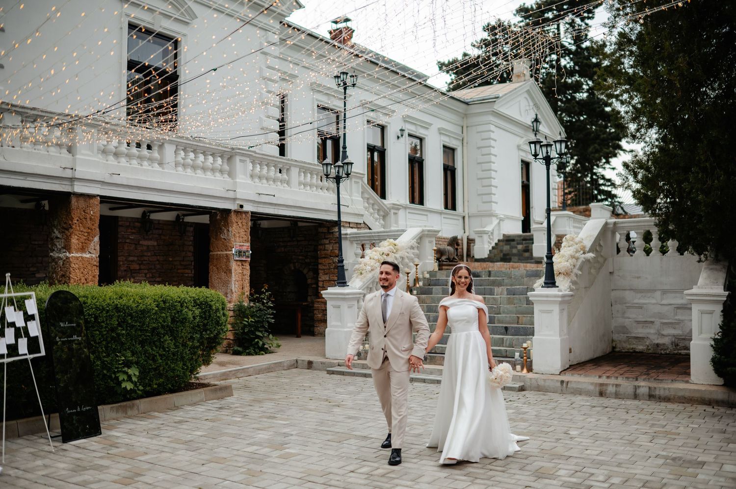 Newlyweds walk joyfully hand in hand outside elegant white wedding venue adorned with string lights and floral decor.