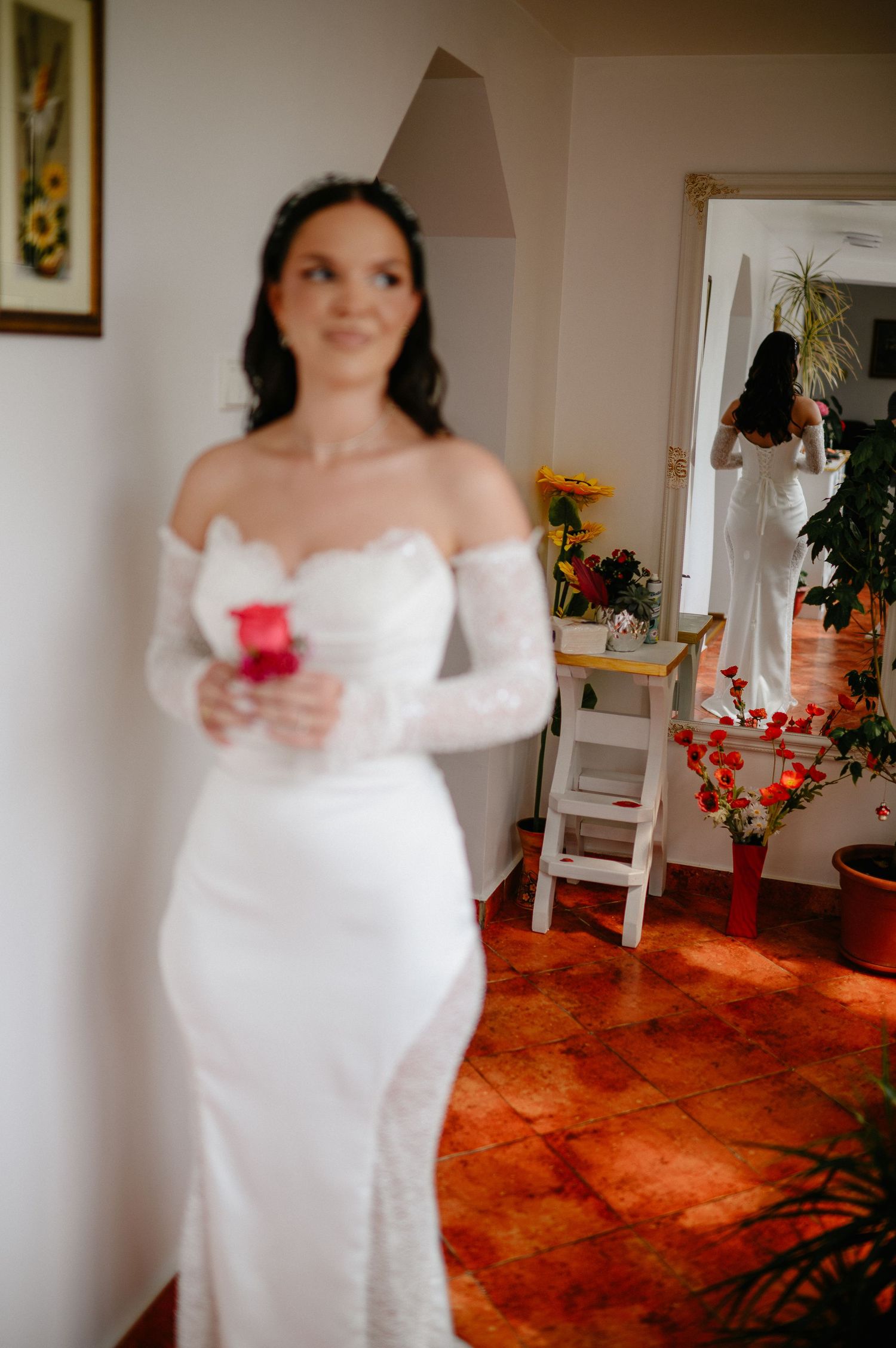 Beautiful bride in fitted white lace wedding dress holding red rose in floral decorated room.