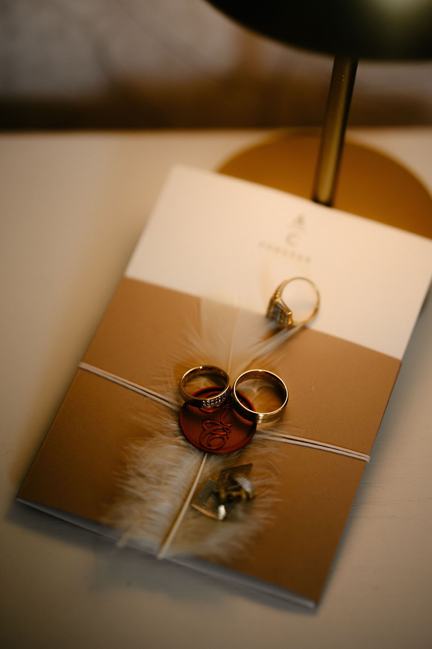 Gold wedding invitation with two rings, wax seal, and white feather on decorated table.